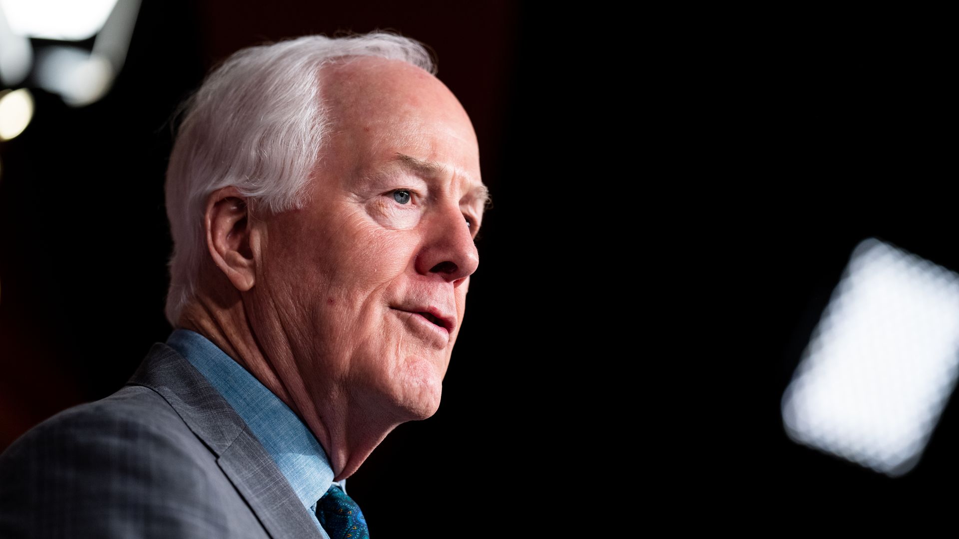 Sen. John Cornyn, under the black ceiling of a TV studio room, wearing a gray suit and blue shirt.