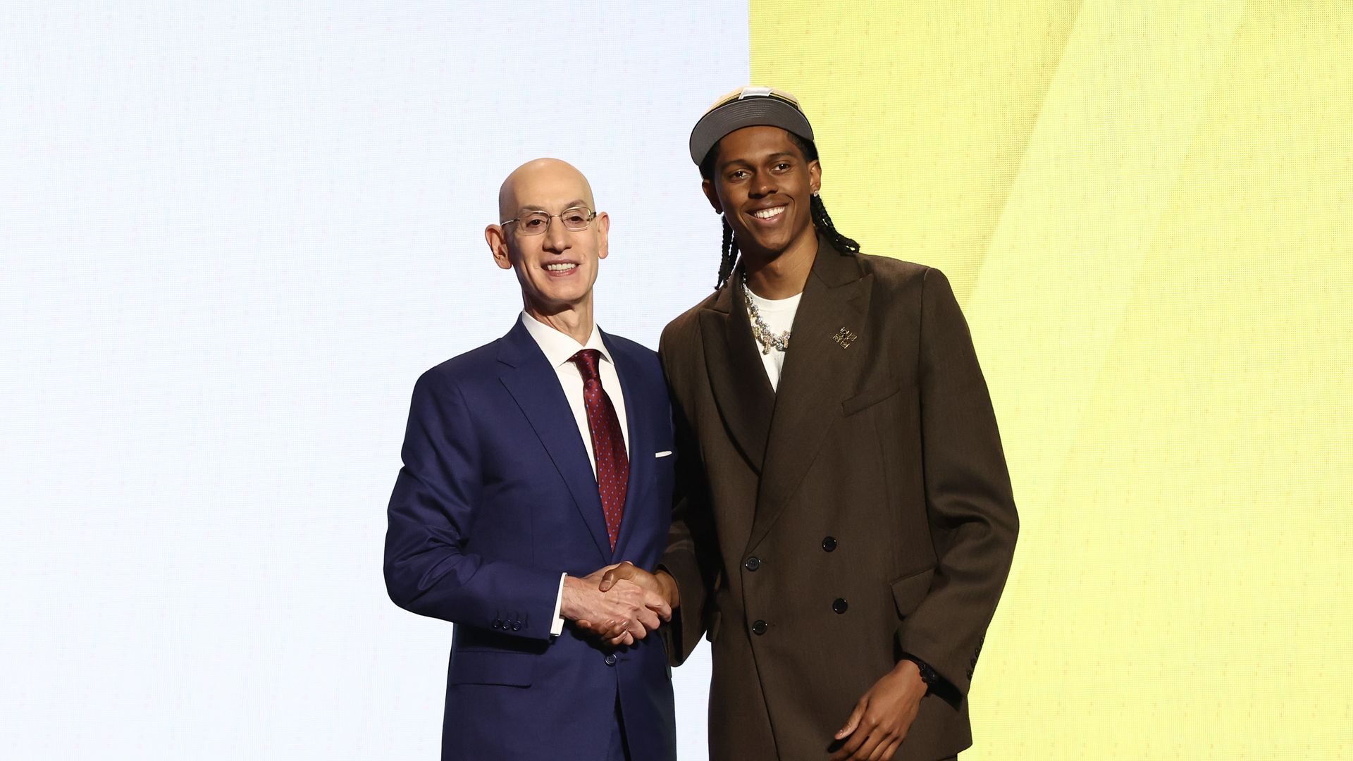 Two men wearing suits shake hands in front of a yellow background at the NBA draft