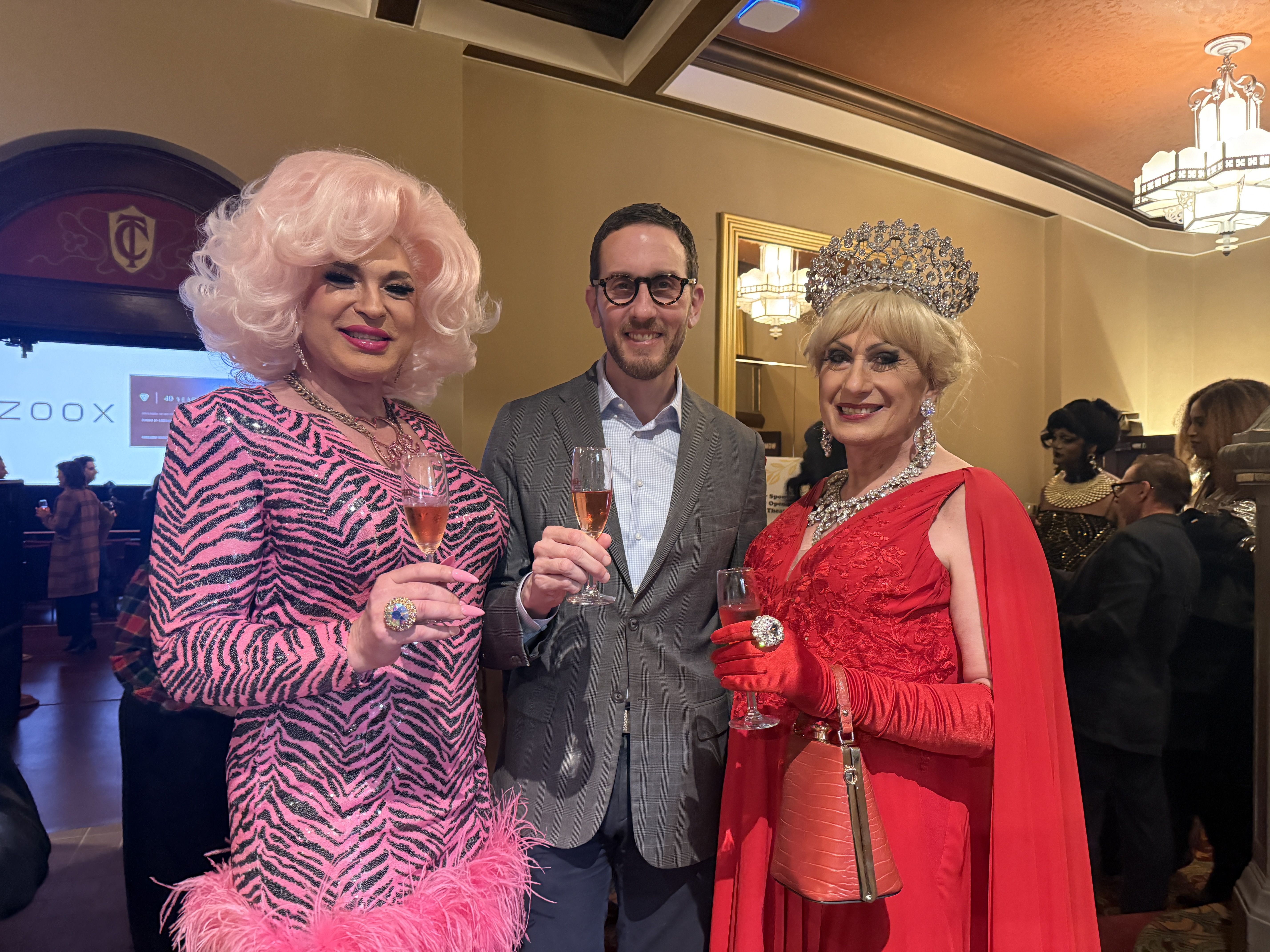 Three people dressed in evening wear raise glasses of champagne inside the lobby of the Castro Theatre with a crowd behind.
