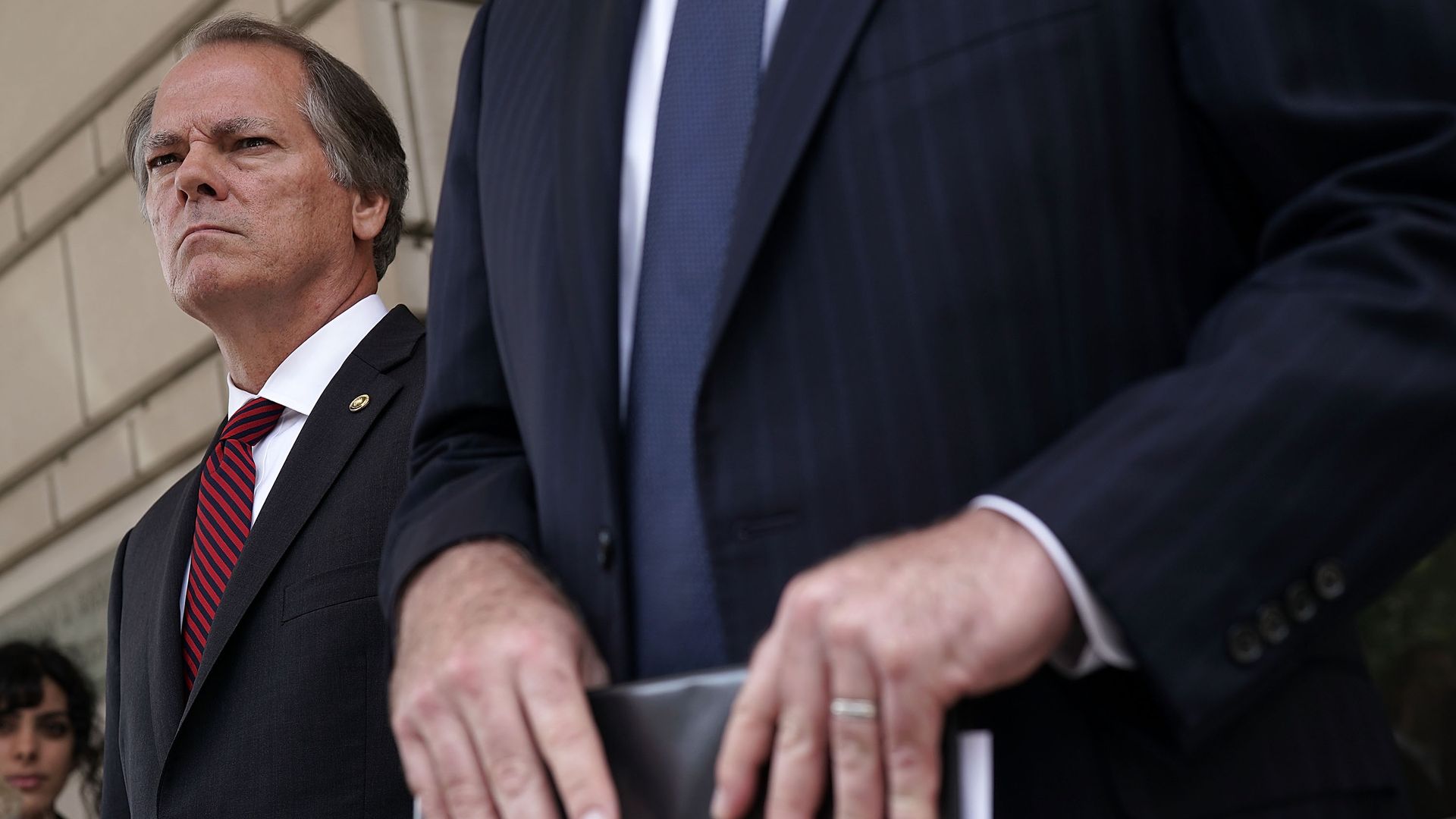 James Wolfe looks with a scowl behind the suit and arms of his lawyer