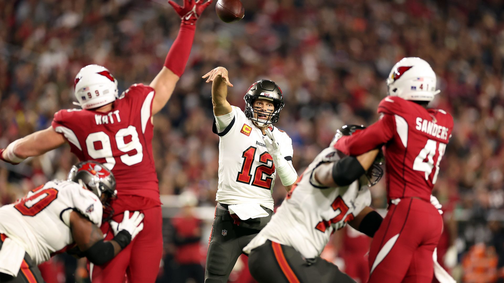 Tom Brady throws a football between two Arizona Cardinals defenders toward the direction of the camera.