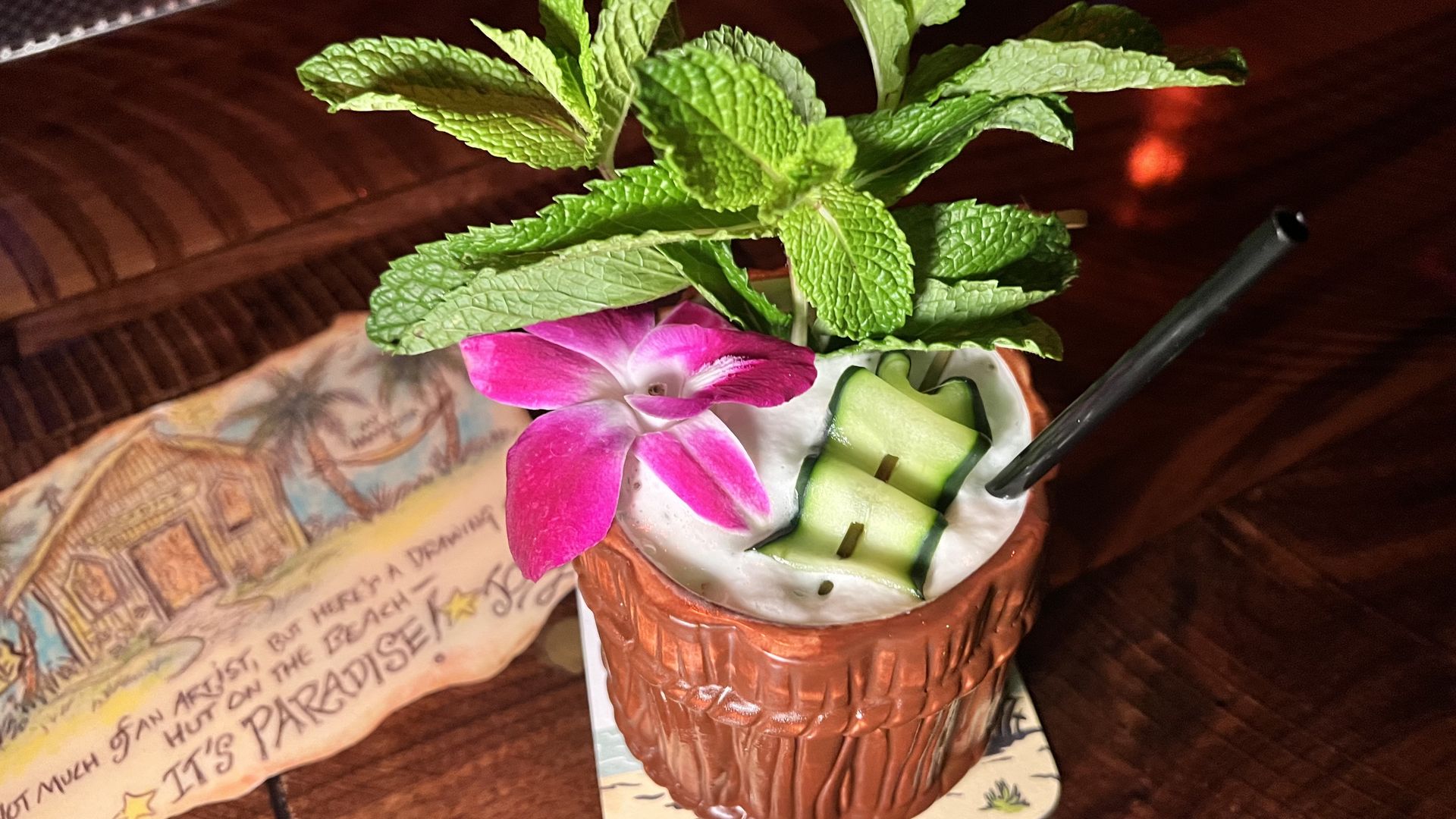 A tropical cocktail served in a ceramic tiki mug topped with white foam, garnished with bright green mint, a pink orchid, and cucumber slices; a straw sticks out as it sits on a wooden table.
