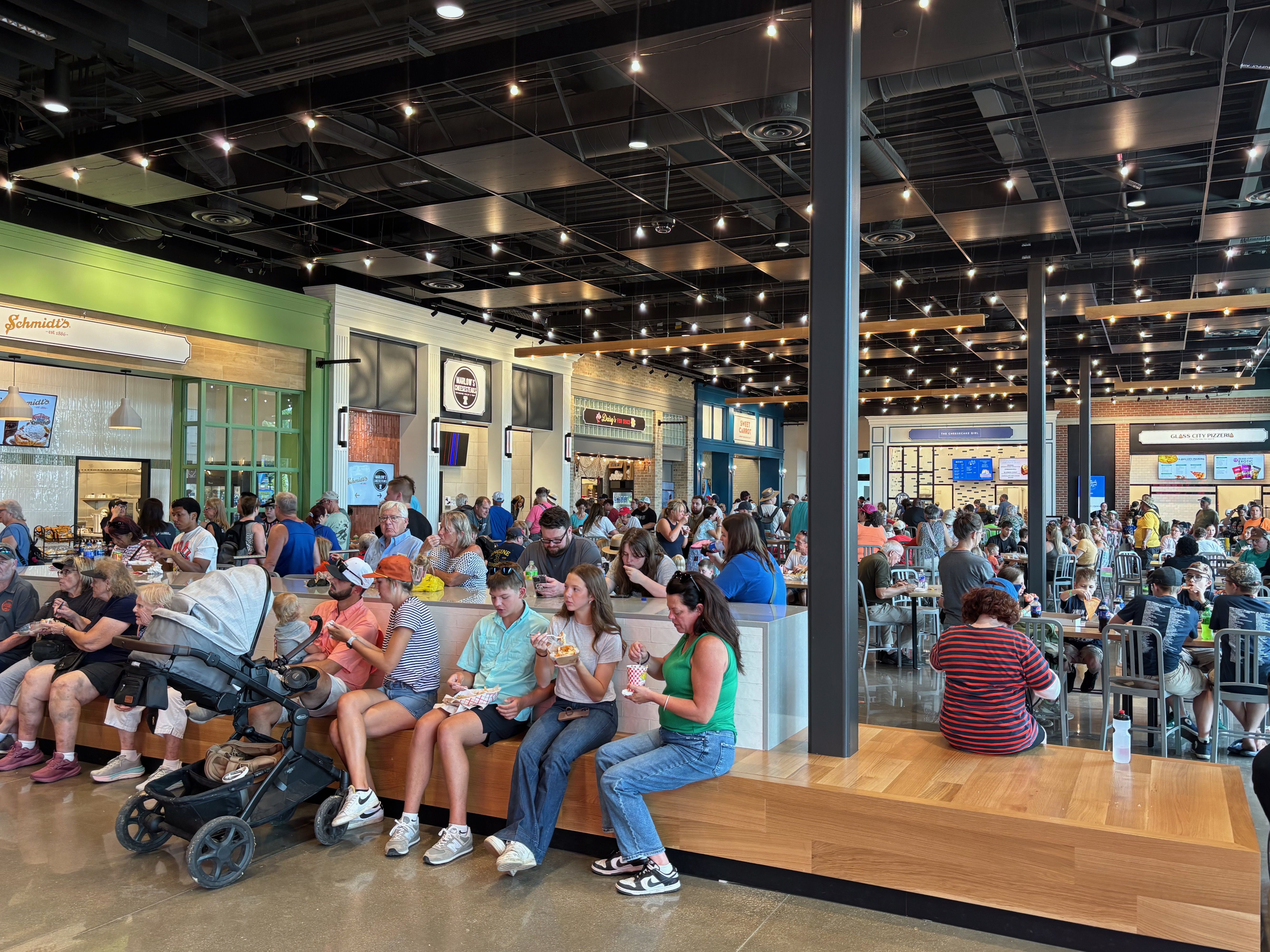 Busy indoor food hall with many people seated at long benches, eating. Exposed ceiling with string lights, storefronts and a green wall on the left, and tall gray pillars in the center.