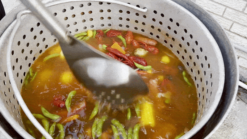 Image shows a giant spoon stirring a pot full of reddish liquid filled with crawfish, corn, sausage, carrots, onions, potatoes and edamame.