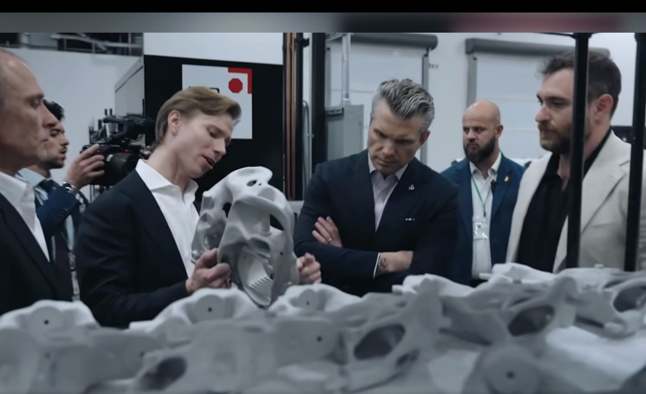 Group of formally dressed men in a bright factory examine a gray, bone-like 3D-printed object; one man holds it while others watch and a cameraman films in the background.