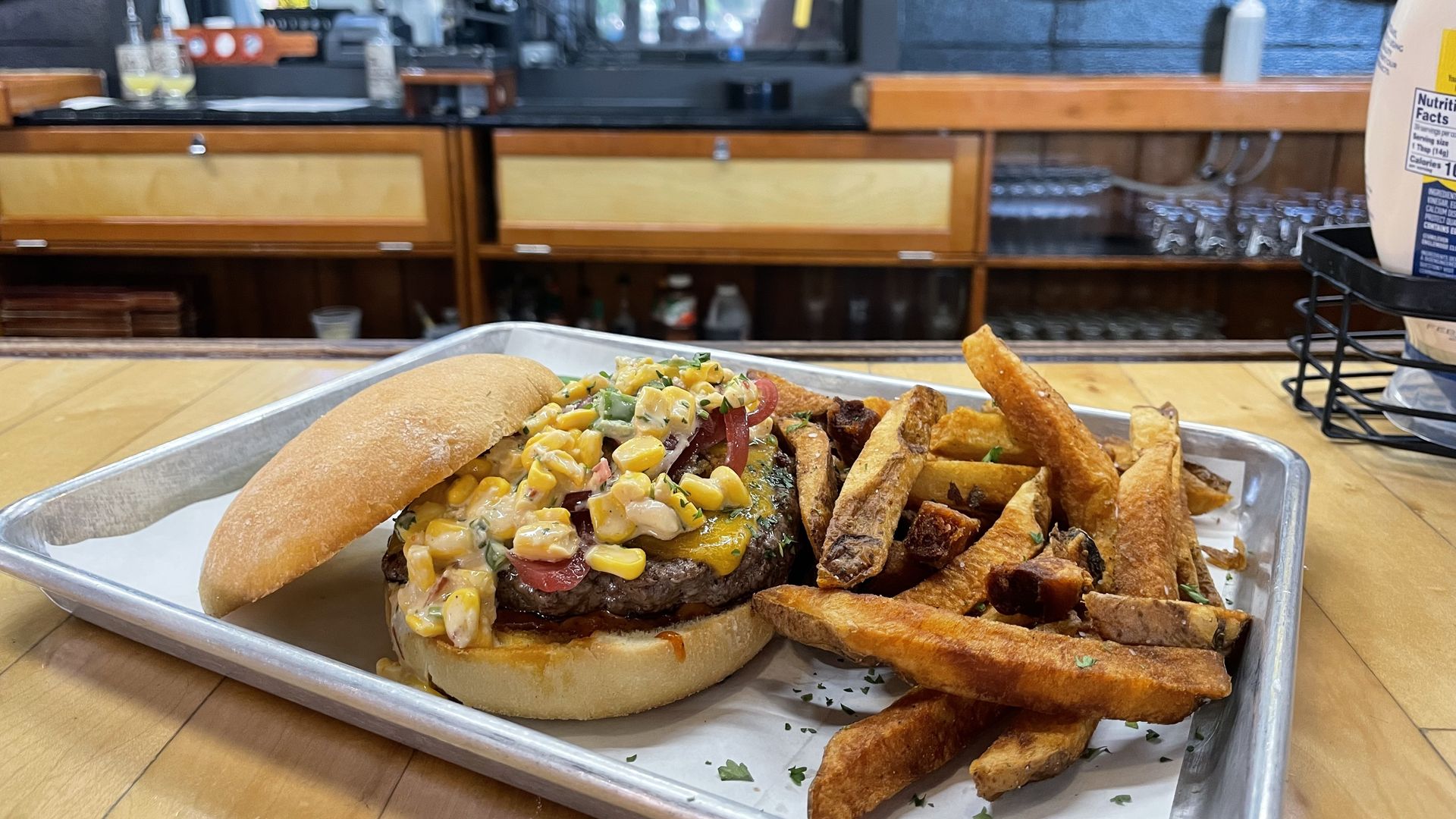 Tray with a loaded burger topped with corn relish, diced peppers, and creamy sauce, plus seasoned fries. Bar counter with wood cabinets and beer taps in the background.