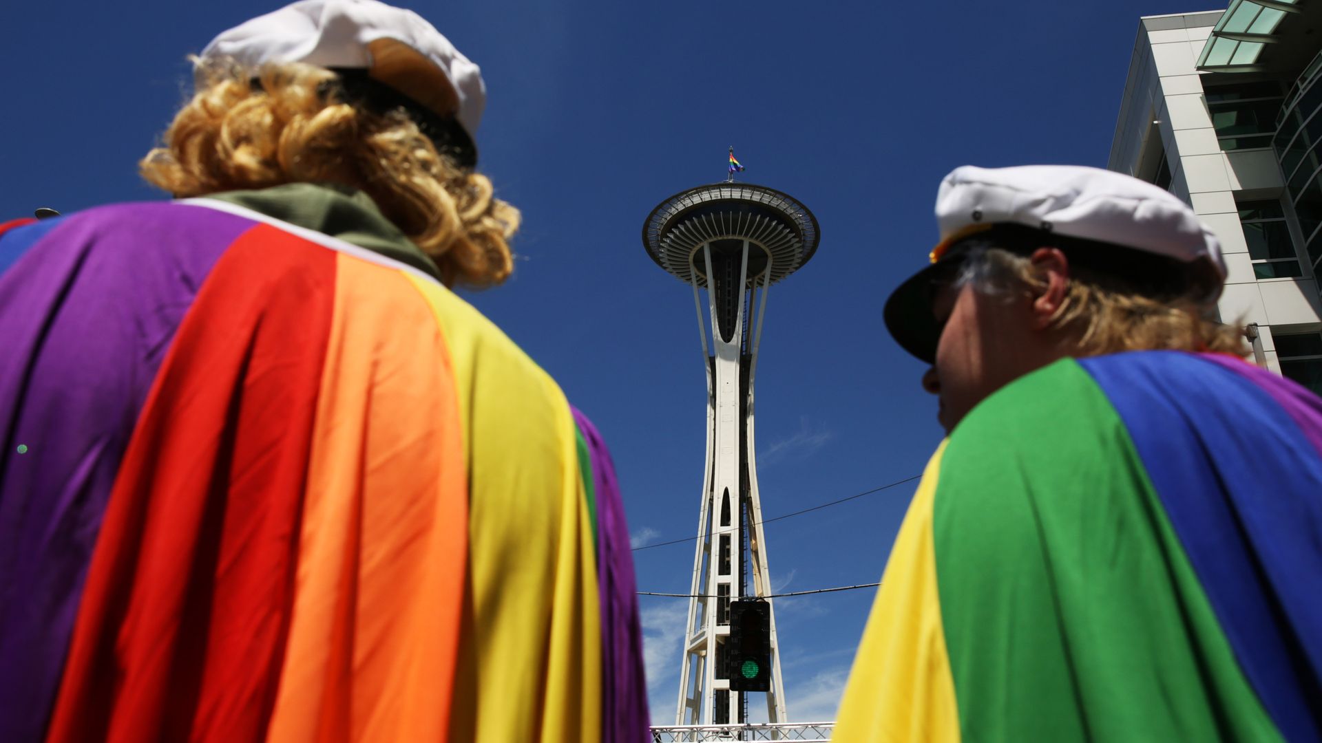 The backs of two people in rainbow cloaks in front of the Space Needle. 
