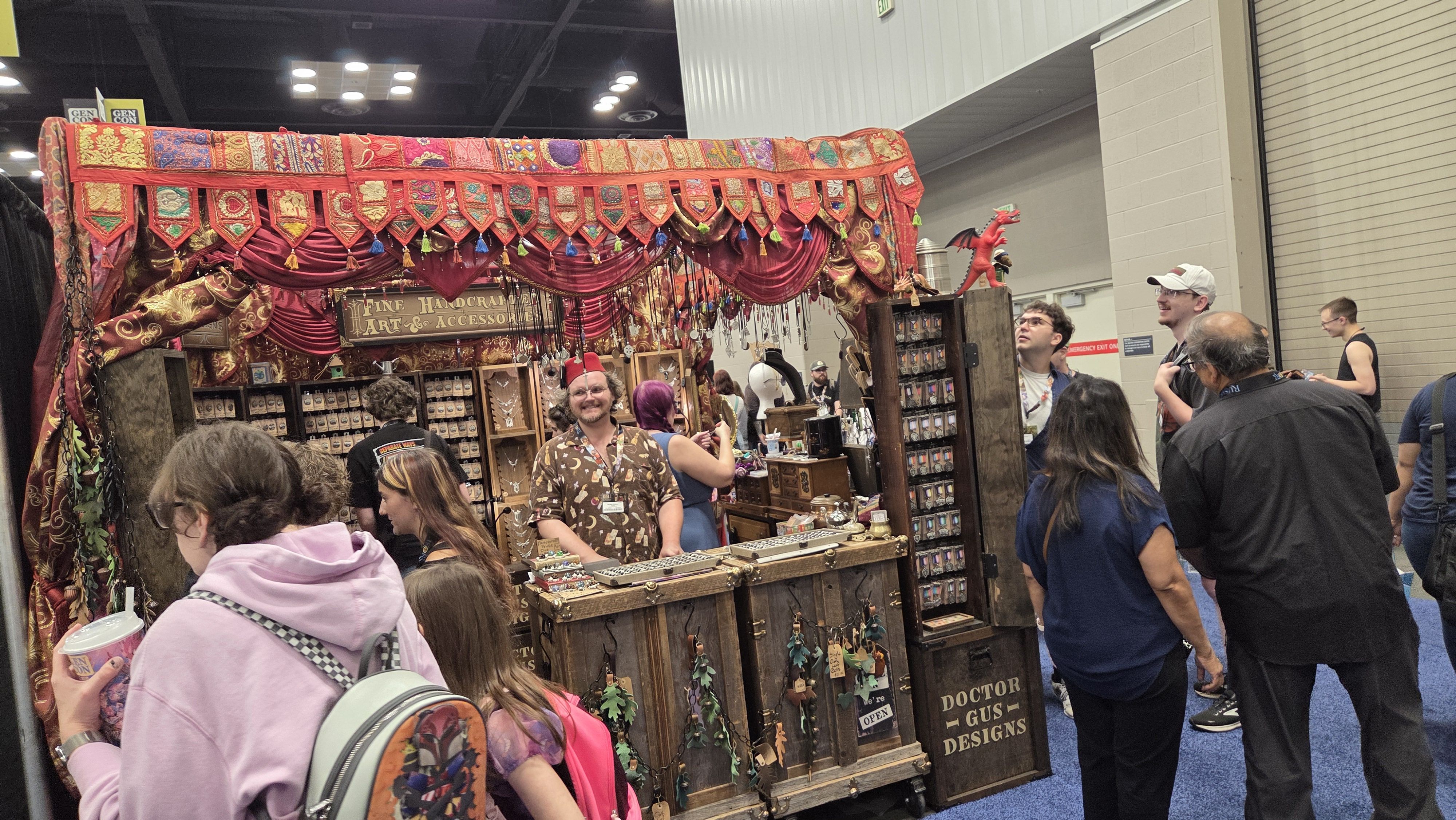 Colorful convention booth with red and gold drapes and tassels, displaying handmade jewelry; a smiling vendor in a patterned shirt chats with attendees near wooden counters.