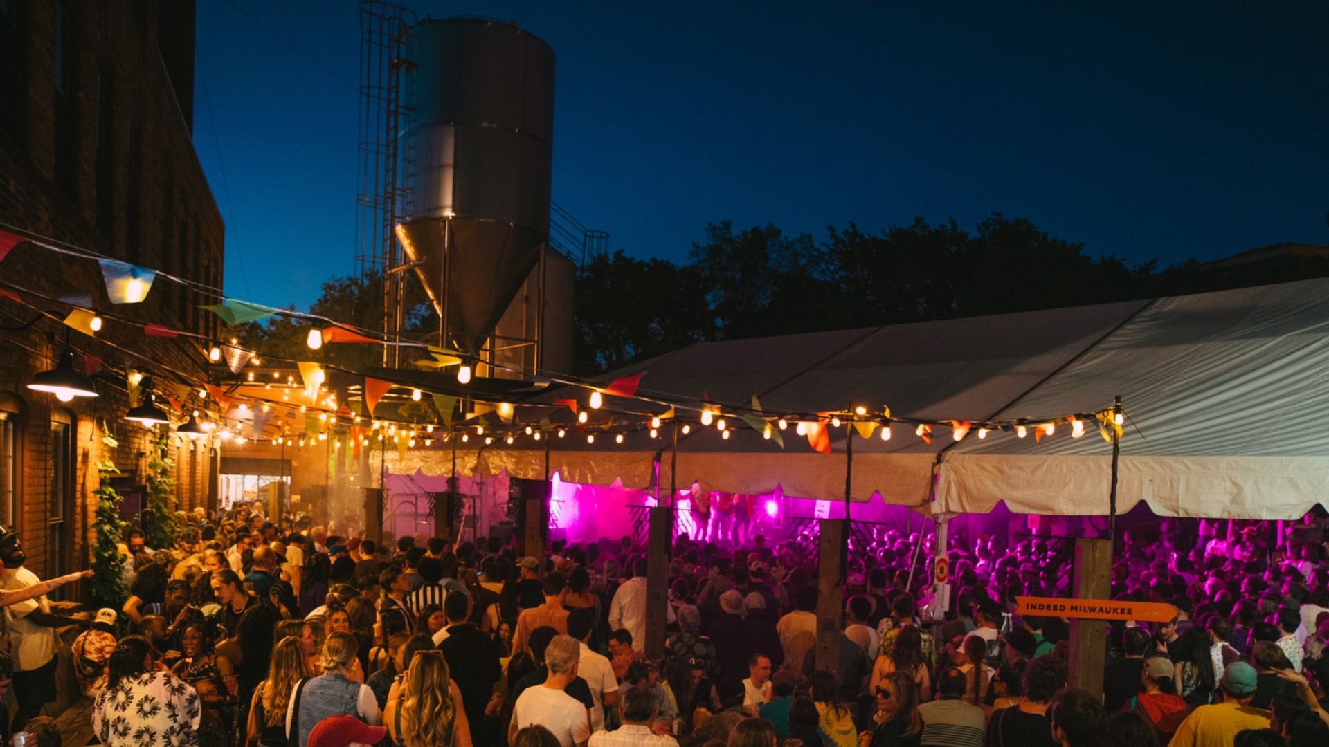 Nighttime street festival with a dense crowd under string lights and colorful pennant bunting; white tents line the area, a stage glows purple in the distance, blue evening sky overhead.