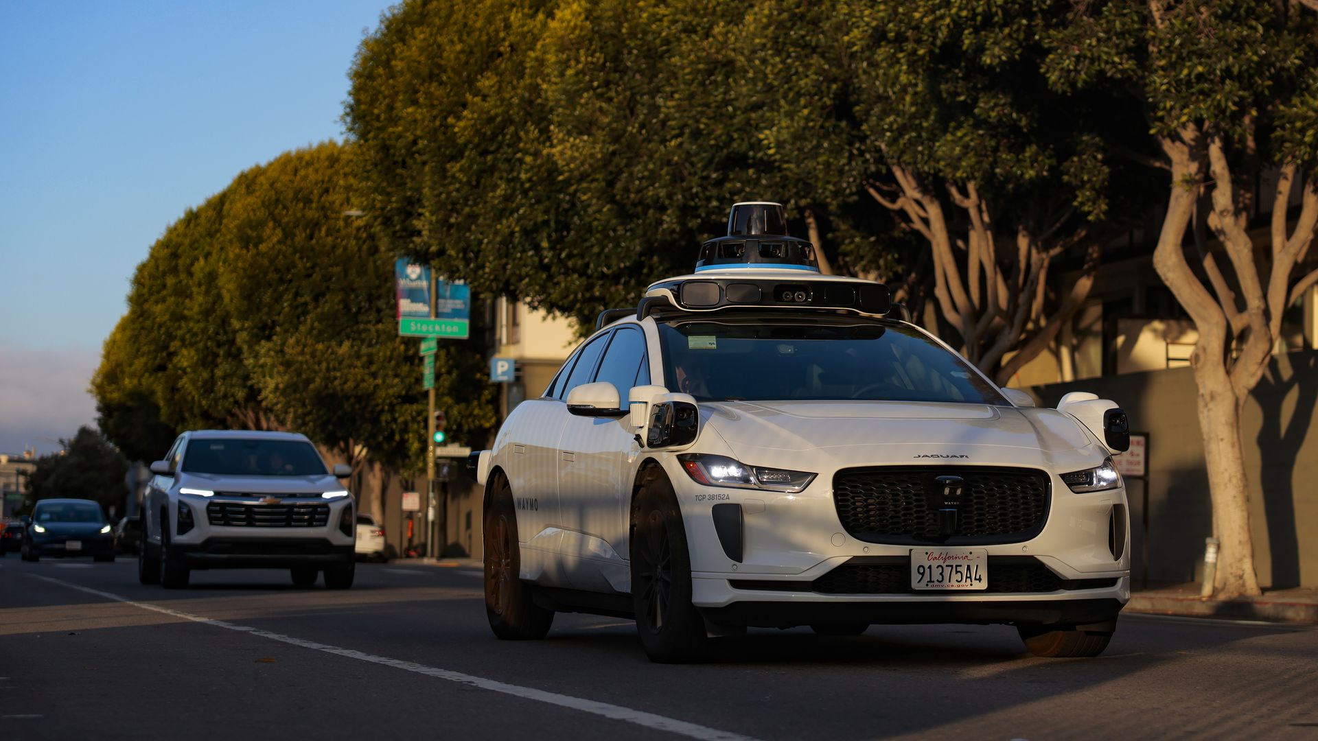 White Jaguar Waymo self-driving SUV with roof sensors in the foreground; California license plate. Street with other cars and tall trees under a clear blue sky.