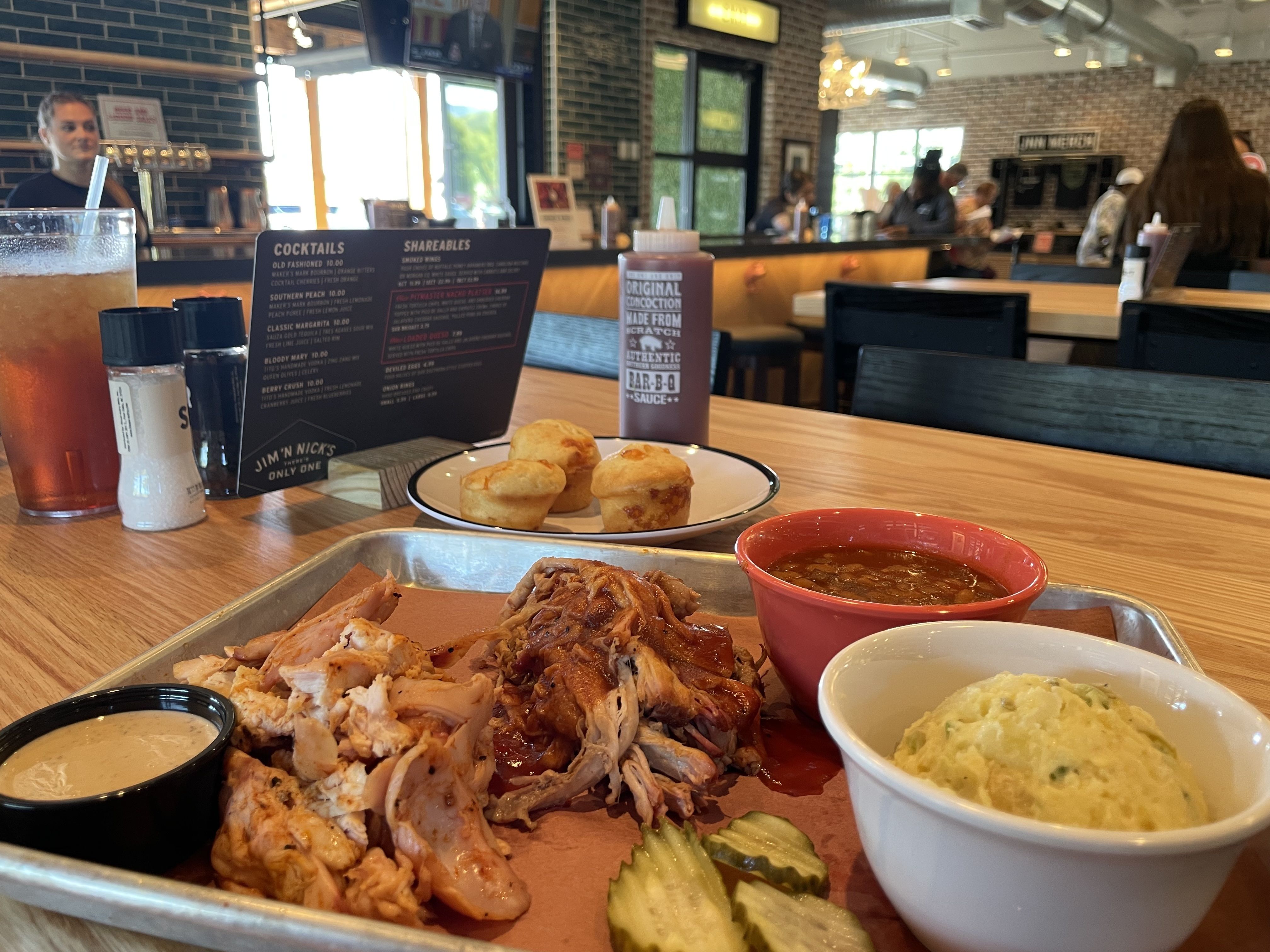 Tray of BBQ food with pulled chicken, pickles, creamy sauce, baked beans, and potato salad on a wooden table with a drink, three cornbread muffins, BBQ sauce bottle, and menu in a casual restaurant.
