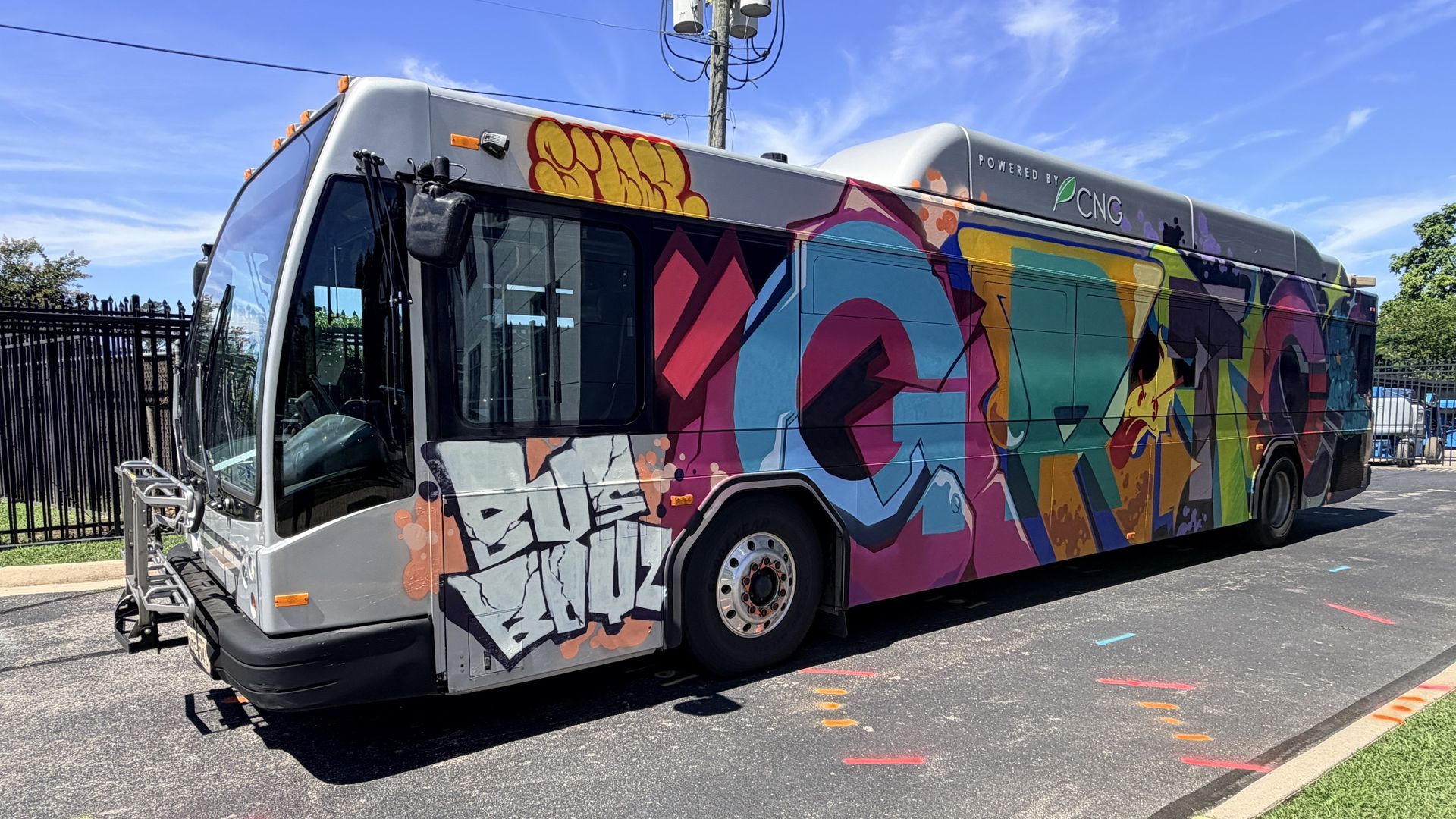 Gray city bus covered in bright graffiti with pink, blue, green, and yellow colors; "POWERED BY CNG" on the roof. Blue sky, power lines, fence, and colorful markings on the asphalt.