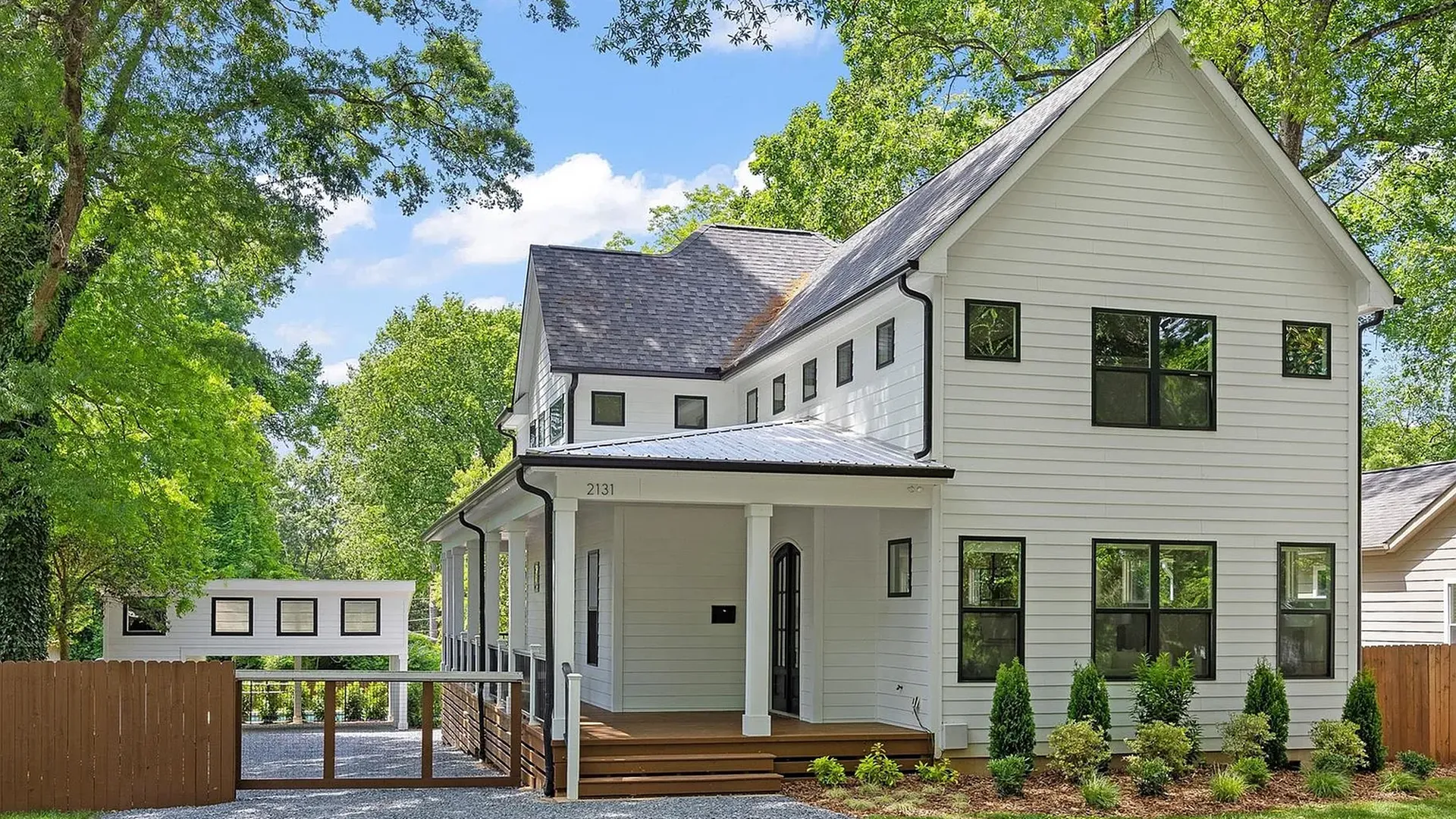 White two-story home with black-framed windows, front porch, and gray roof, amid lush green trees. Gravel driveway to a wooden gate; a small white outbuilding and shrubs in front.