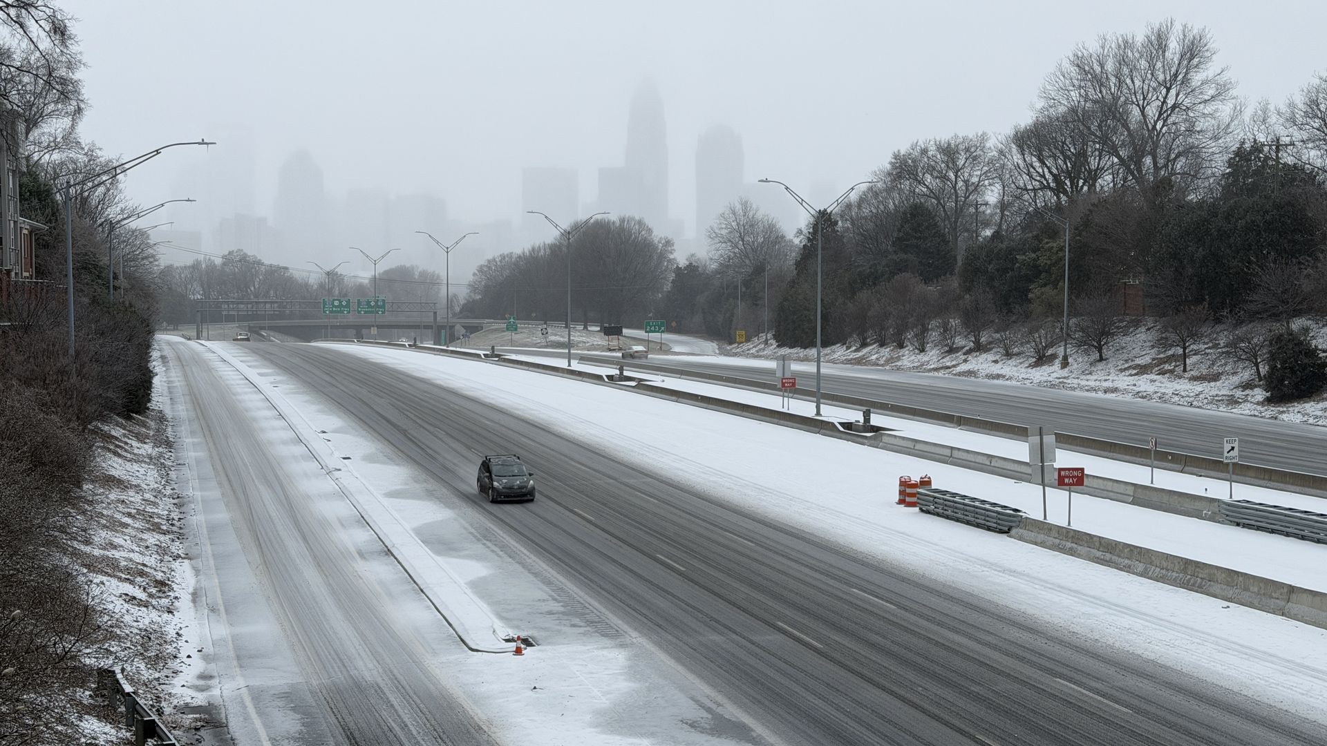 Snow-covered highway with a single black car driving; foggy city skyline in the background; leafless trees and bushes line the road; overcast sky with muted colors.