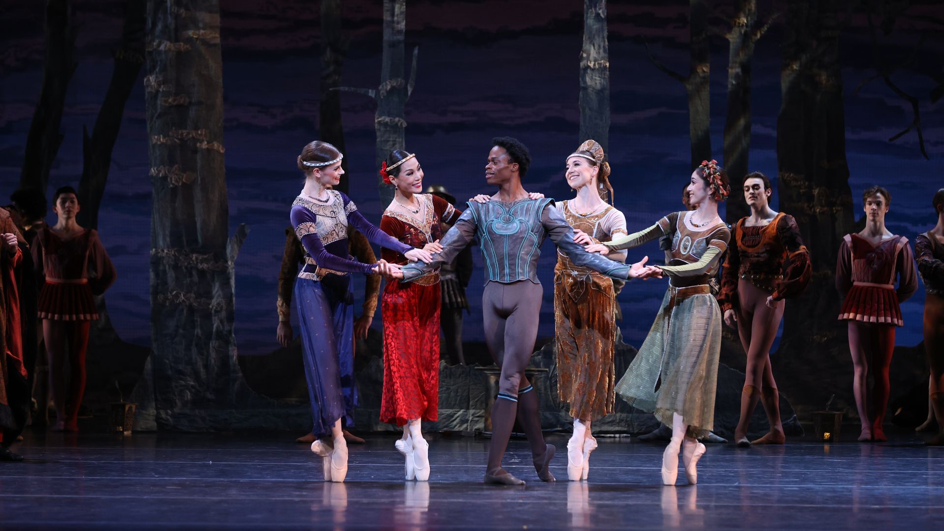 Group of ballet dancers in ornate period costumes form a circle, holding hands on a dark stage with tall tree props and a dusky forest backdrop, surrounding a central dancer in blue-gray.