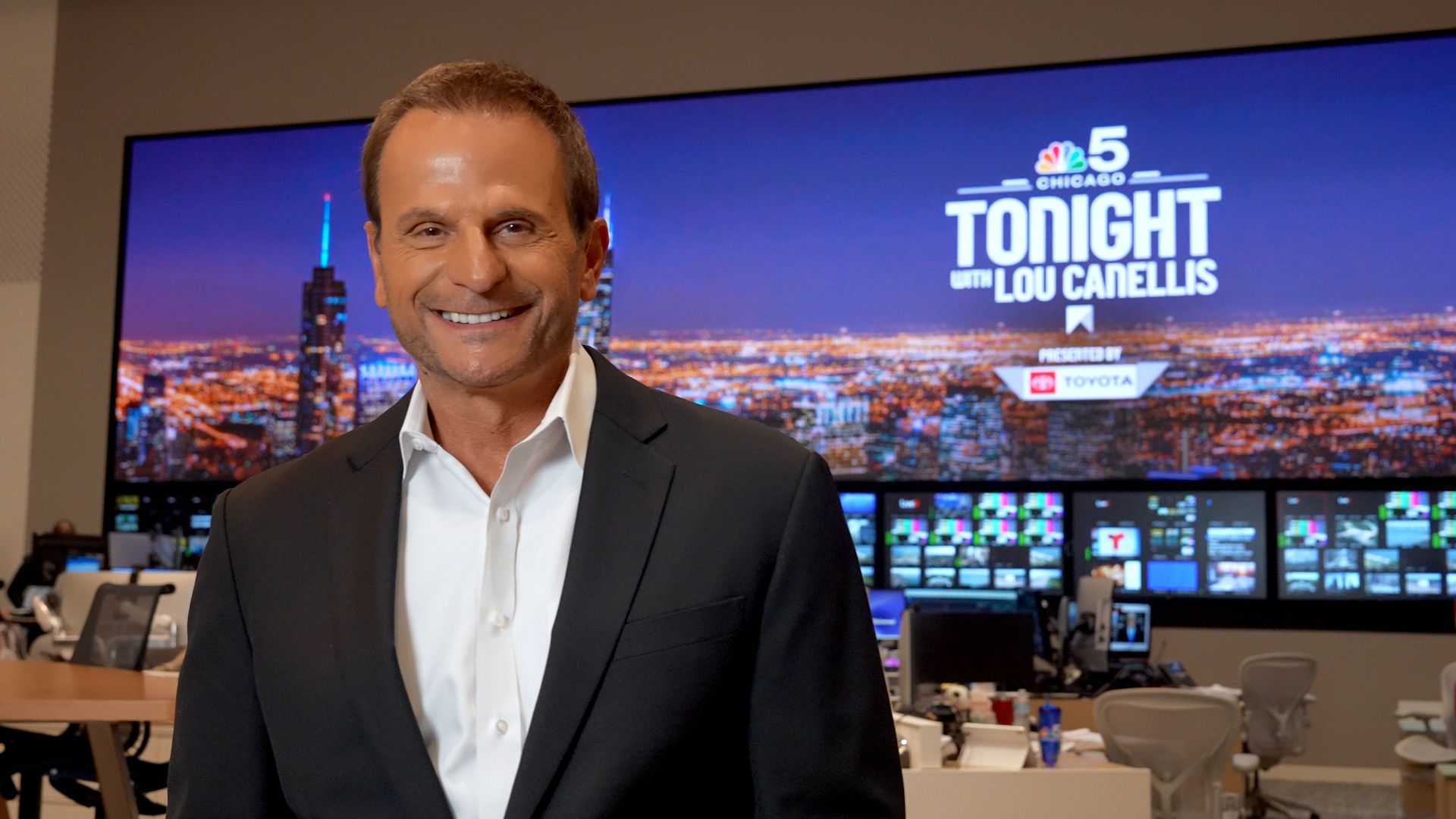 Smiling man in a dark suit and white shirt stands in a newsroom. Behind him, a large screen displays city lights and the title "Tonight with Lou Canellis" alongside NBC 5 Chicago branding.