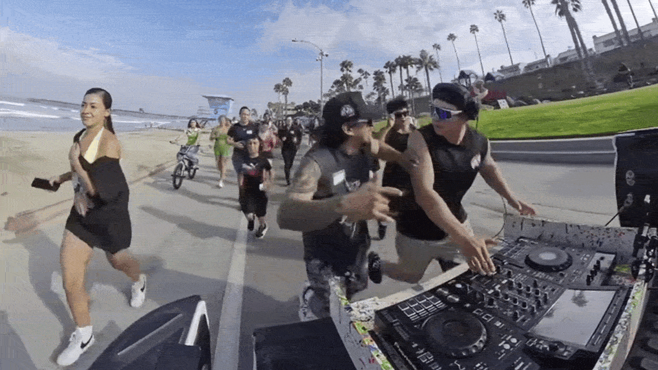 Beachside scene with a DJ at turntables on the right, a crowd running and dancing along a coastal promenade, palm trees in the background and the ocean on the left.