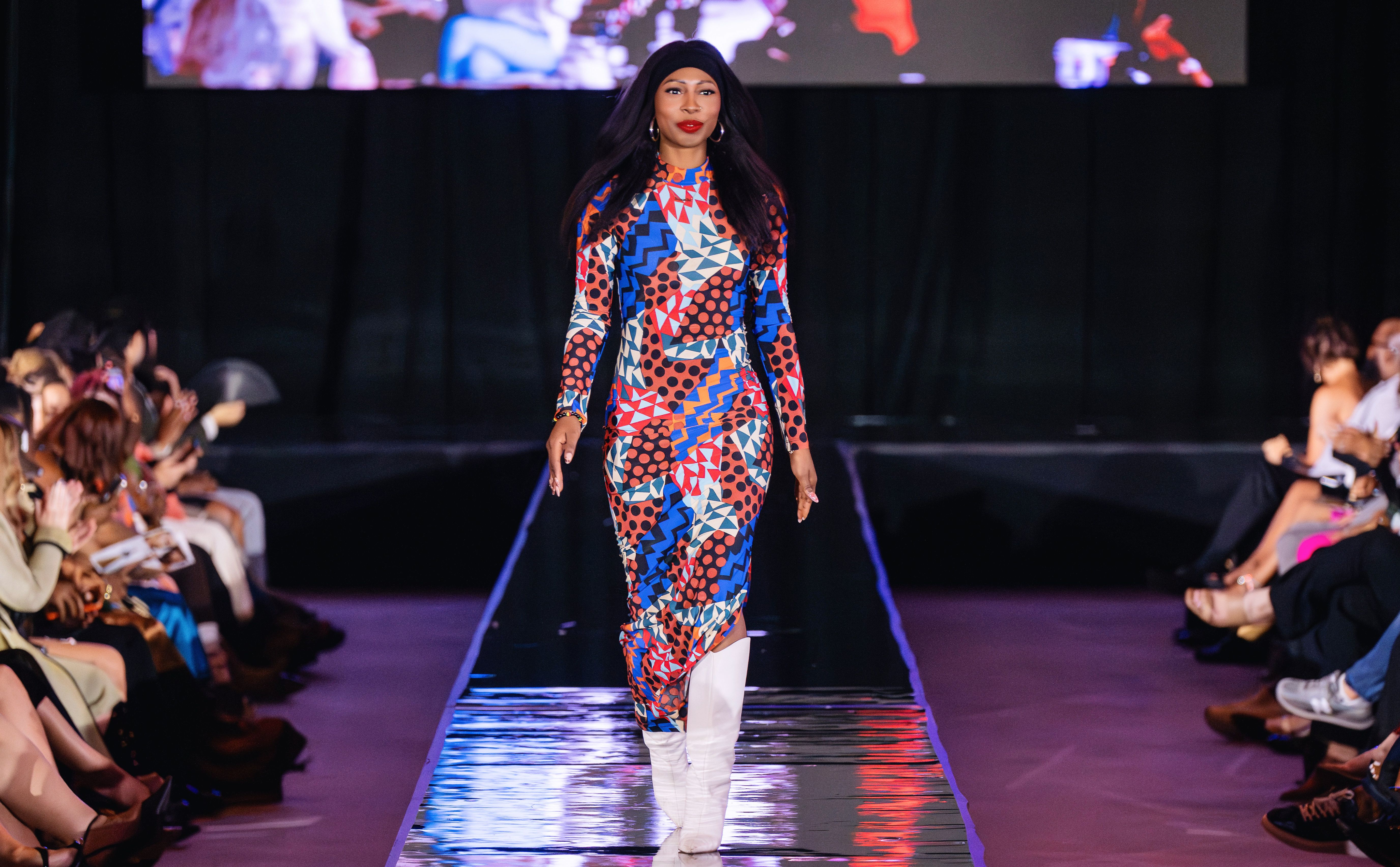 Model walking on runway wearing a colorful geometric and polka dot patterned dress with blue, red, white, black, and orange. She has long dark hair and white boots. Audience claps on sides.
