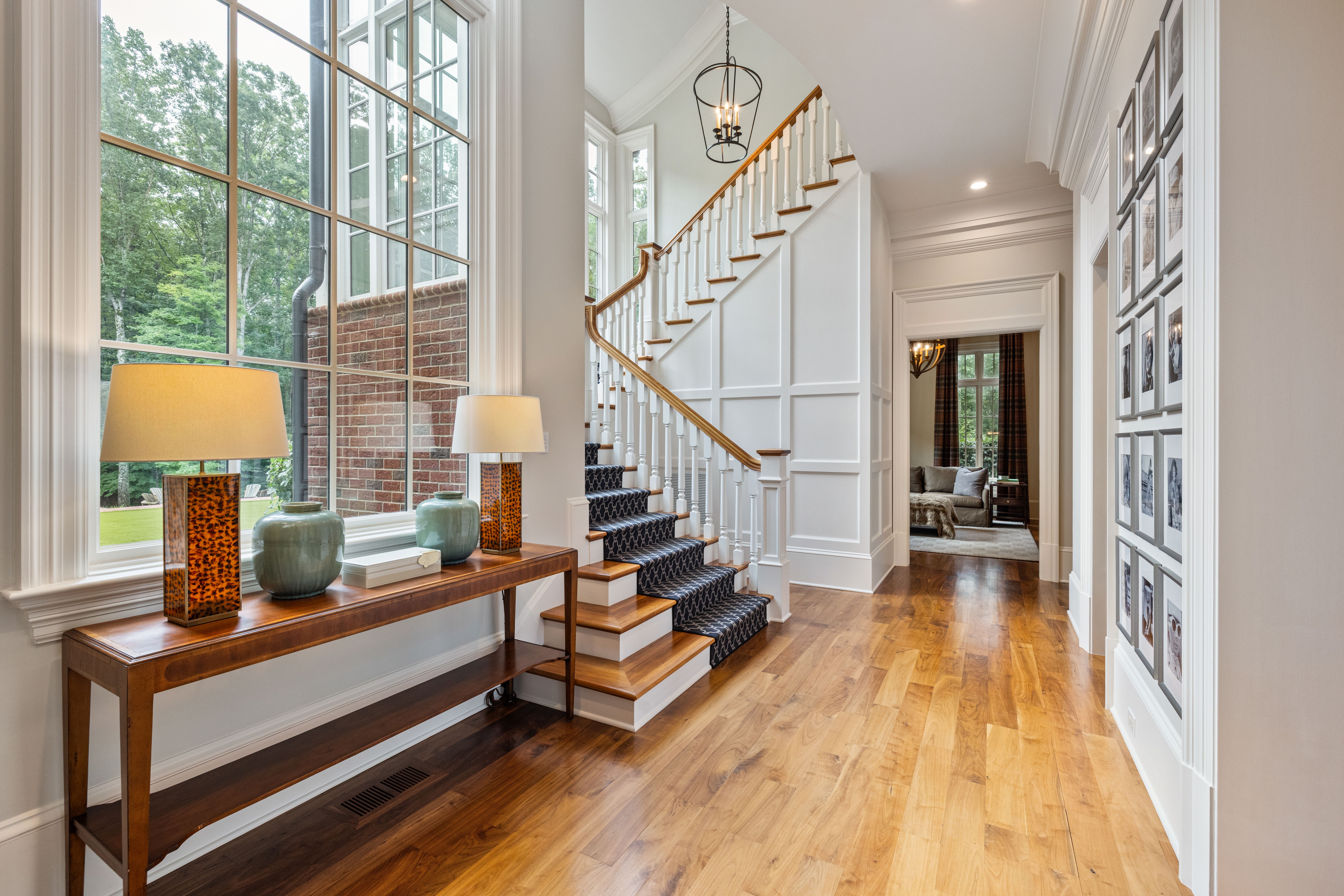 Bright hallway with wooden floor, white staircase with blue patterned runner, wooden console table holding two lamps and green vases, large window showing outdoor greenery, photo gallery wall.