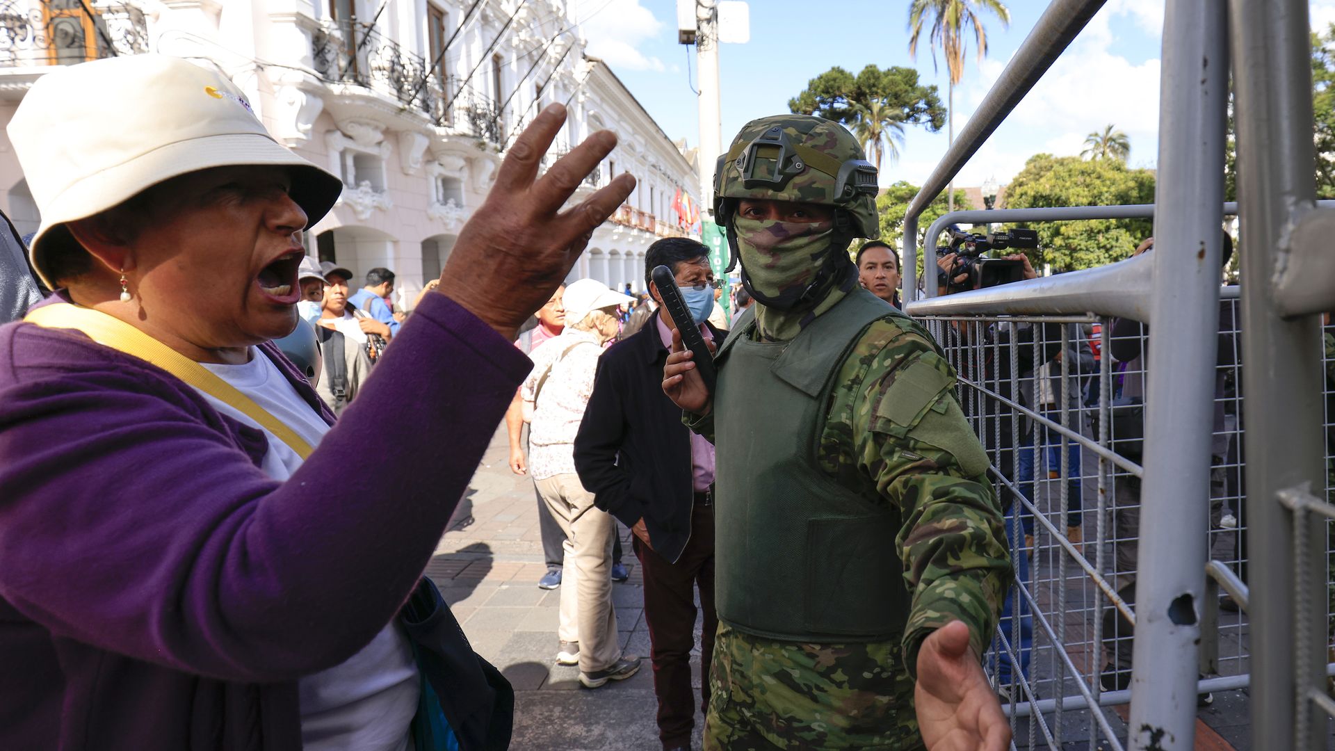 A woman wearing a bucket hat has her arm out and appears to be yelling next to a soldier in uniform outside government palace in Quito, Ecuador