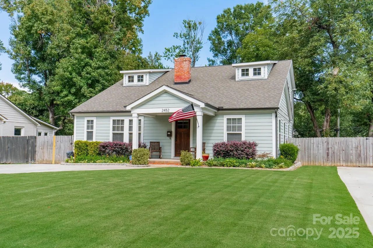 Light gray single-story house with white trim, American flag by wooden front door, green lawn, shrubs, and tall trees behind wooden fence under blue sky.
