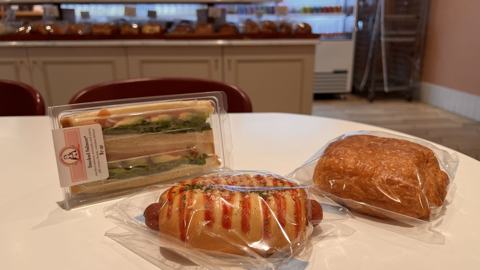 Table with three packaged bakery items: a smoked salmon sandwich with lettuce, a hot dog bun with ketchup drizzle, and a rectangular pastry, all in clear plastic wrap.
