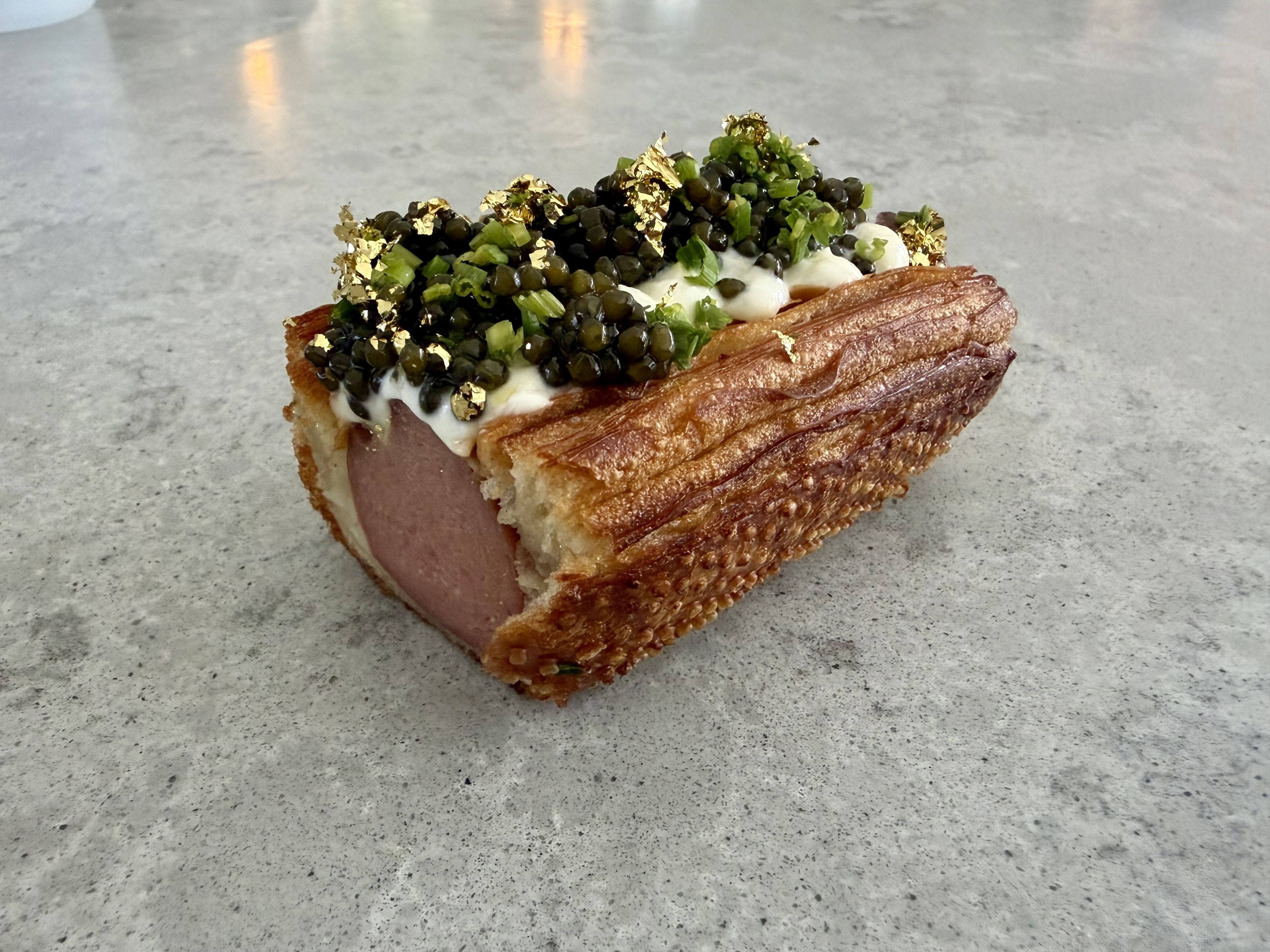 Hot dog with toasted bun, topped with white sauce, black caviar, green herbs, and gold flakes, placed on a gray textured surface.