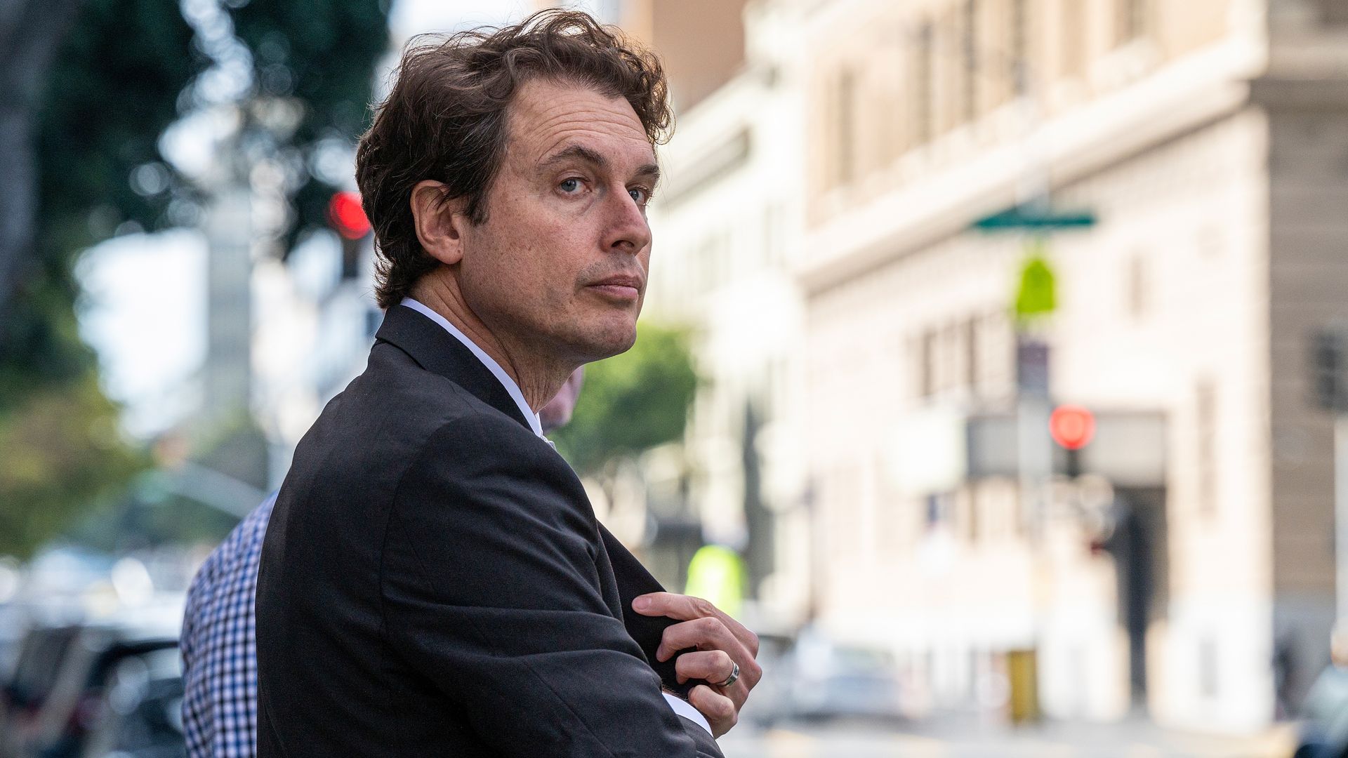 Kimball Musk in a black suit with curly brown hair, standing on a city street looking sideways, holding his jacket. Background shows blurred buildings, cars, and trees.