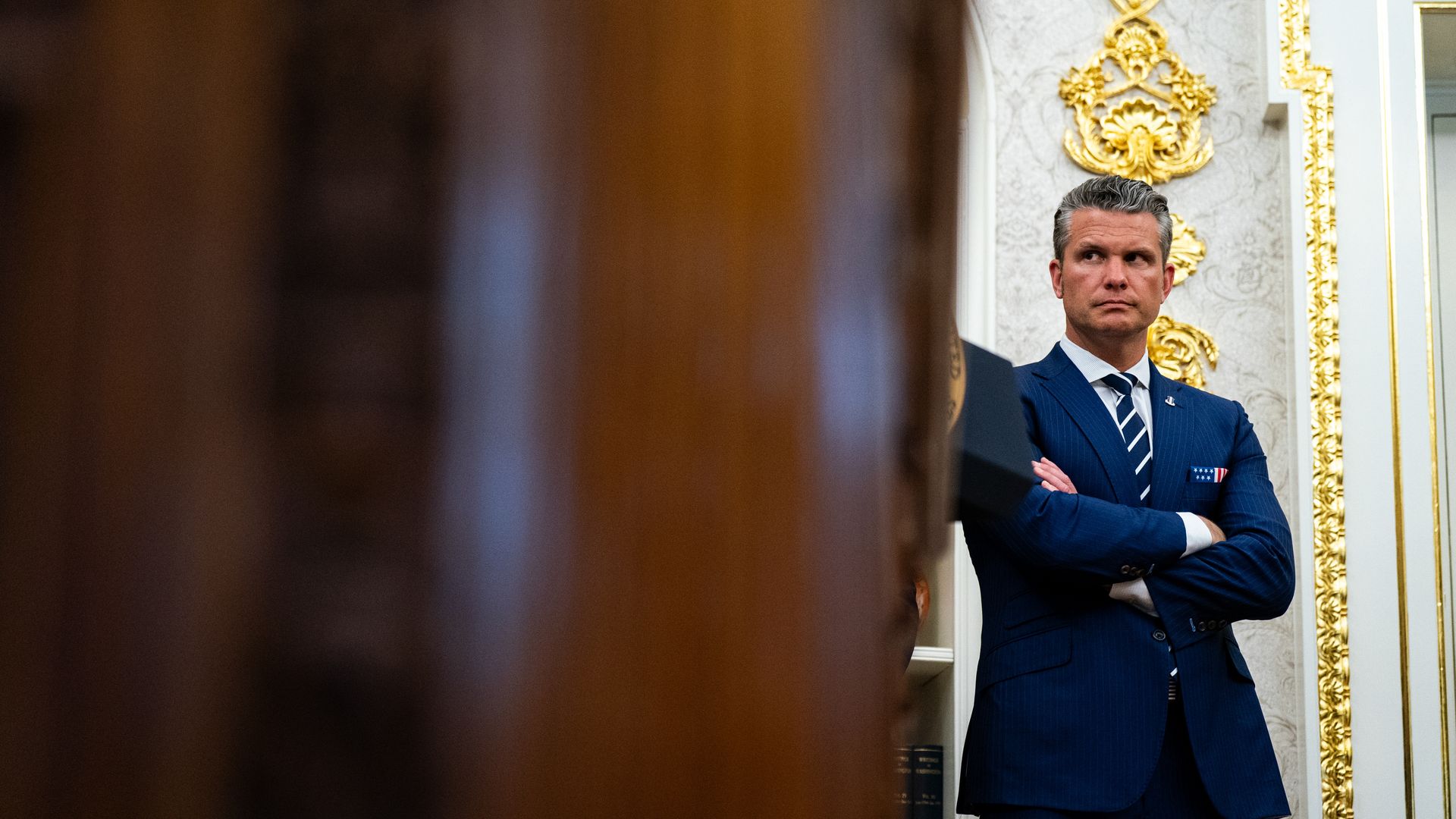 Pete Hegseth, a man, wears a blue suit and blue tie. He is standing in the Oval Office.