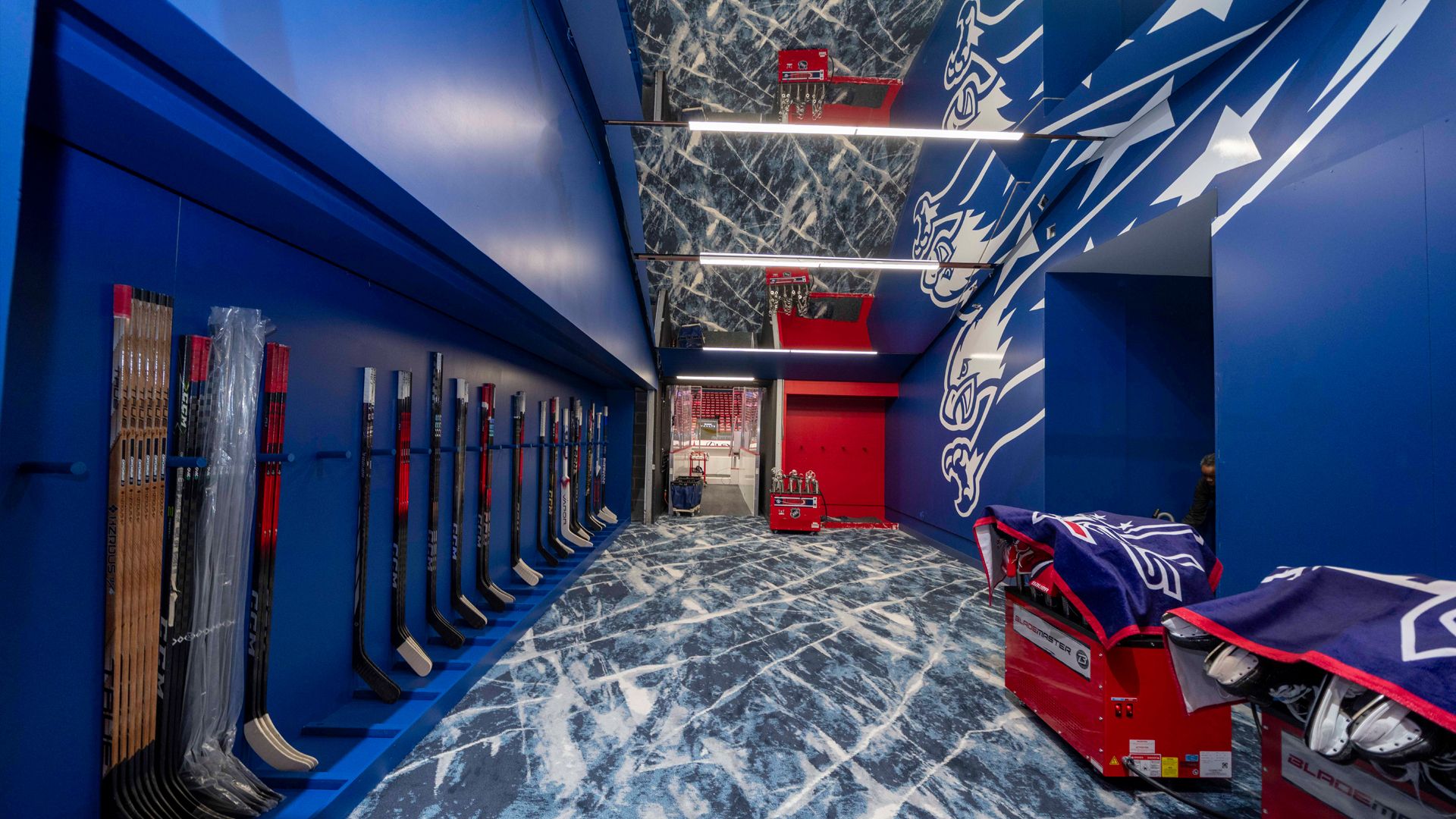 Blue-walled hockey locker room with hockey sticks lined on left wall, a floor with ice-scratch design, and benches on right topped with blue towels featuring a white eagle logo.