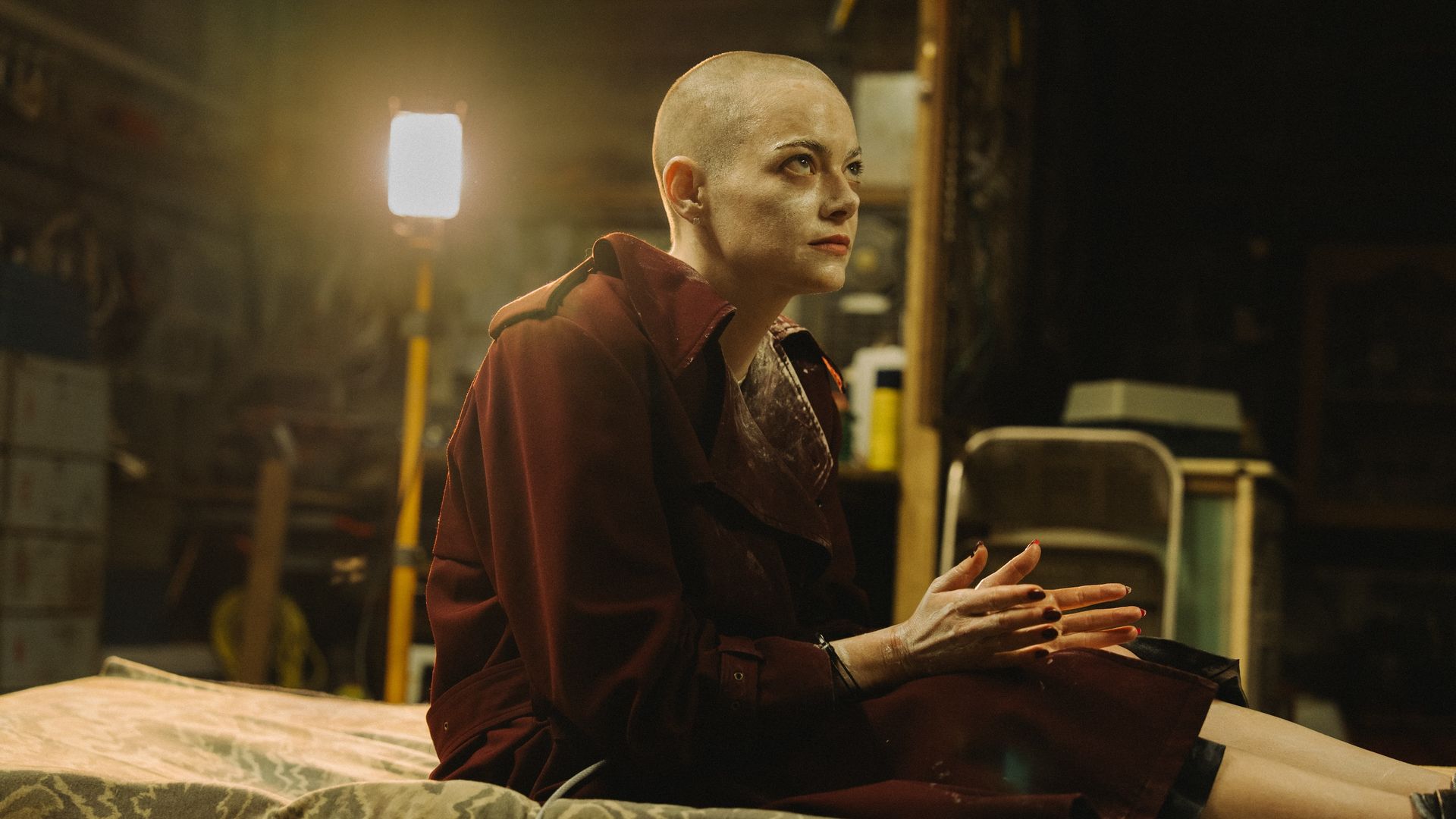 A bald person with intense gaze, wearing a dark red coat, sits on a mattress in a dimly lit room with industrial and cluttered background.