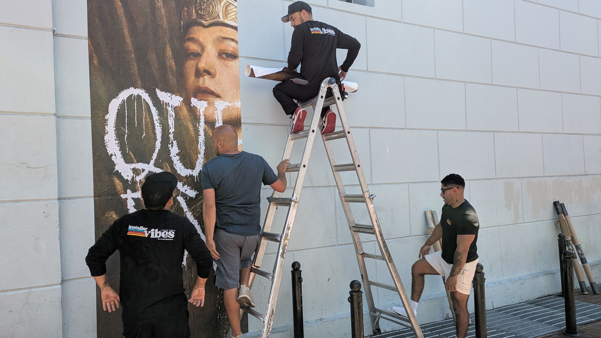 Multiple workers on ladders install a large mural of a crowned "Mona Lisa" on a light-gray exterior wall; a window is above and several onlookers watch.