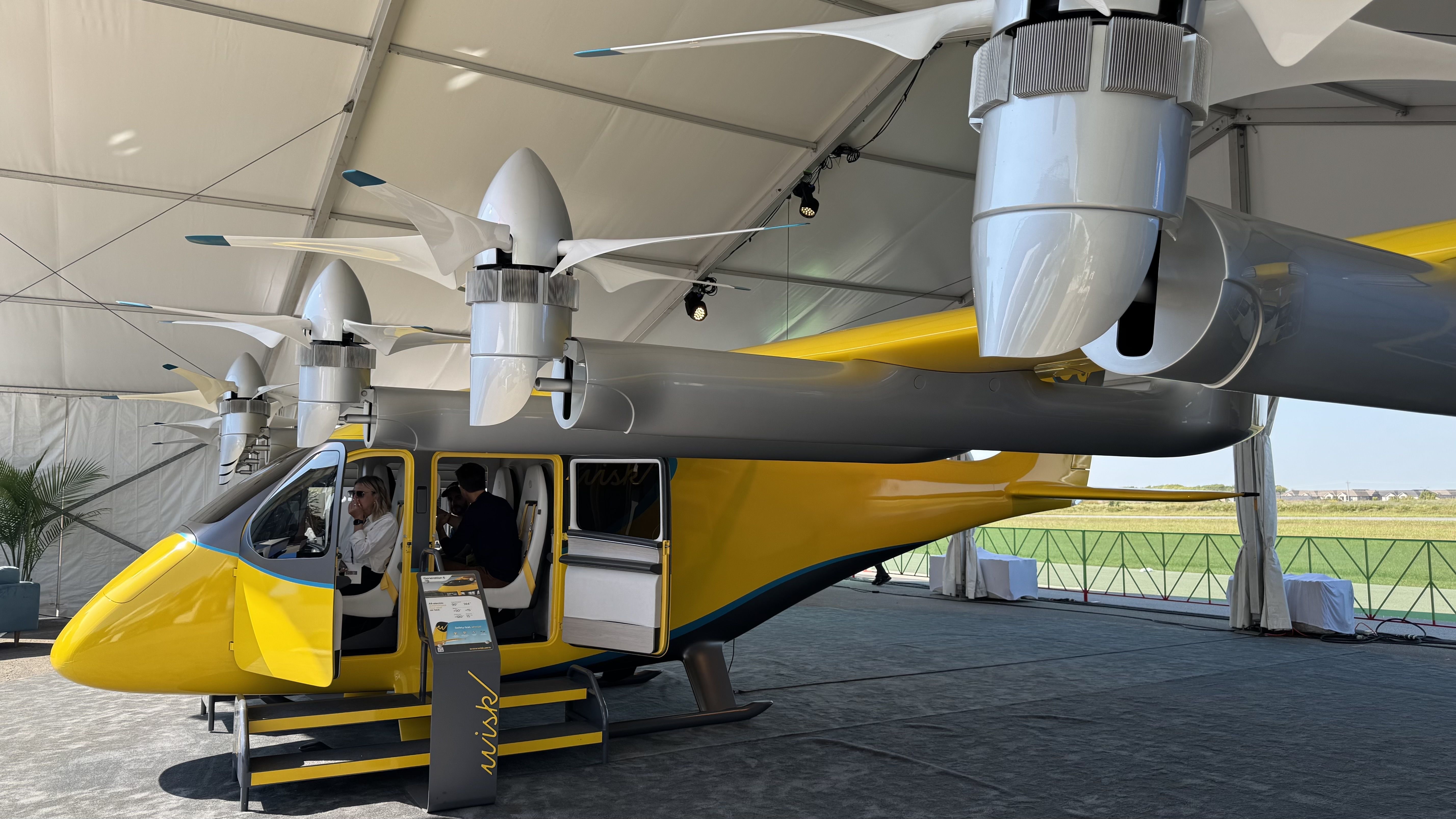 A yellow and gray electric vertical takeoff aircraft with six rotors is on display under a tent. Two people sit inside the cockpit of the sleek, futuristic vehicle, branded with the name "Wisk." The aircraft is showcased on a carpeted floor with a field visible outside the open tent.