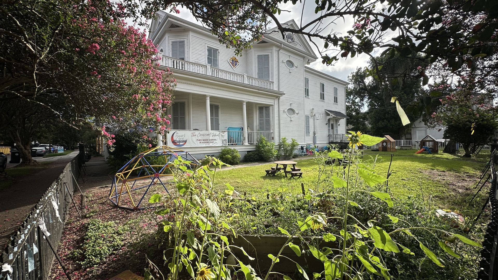 White two-story building with porch and banner reading The Crescent School, surrounded by greenery, bright yellow sunflowers, pink flowering trees, playground structures, and a picnic table.