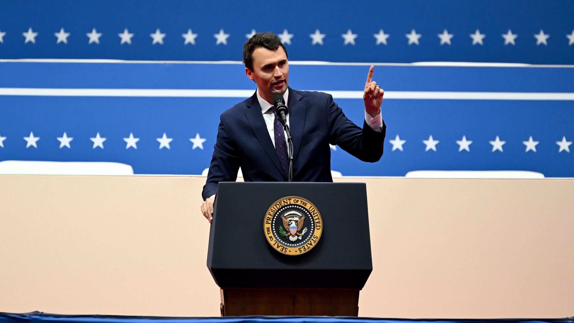 The late Charlie Kirk, founder and executive director of Turning Point USA, points as he speaks at a podium with the words "President of the United States" on it in front of the stars and blue background of the Stars and Stripes US flag.