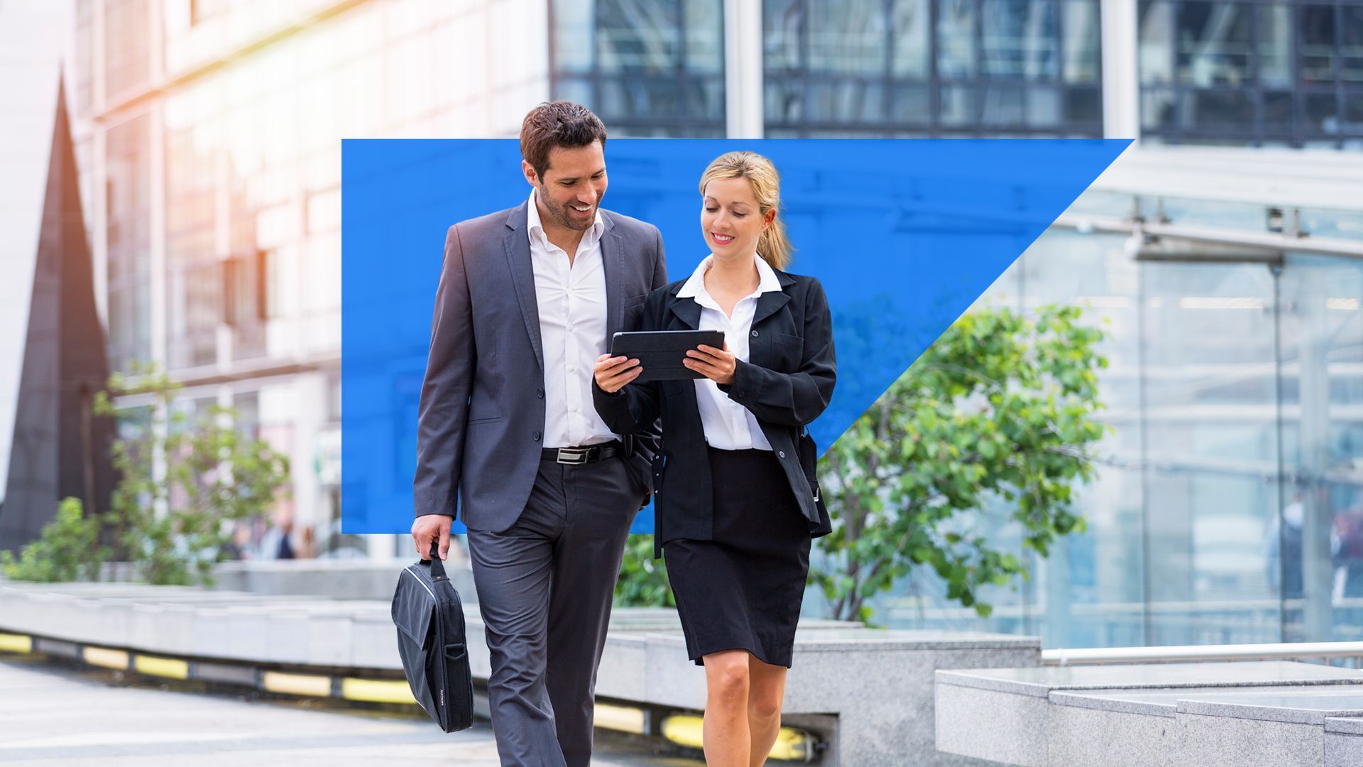 Two business professionals, a man in a gray suit and a woman in a black skirt suit, walk outside an office building while looking at a tablet together.