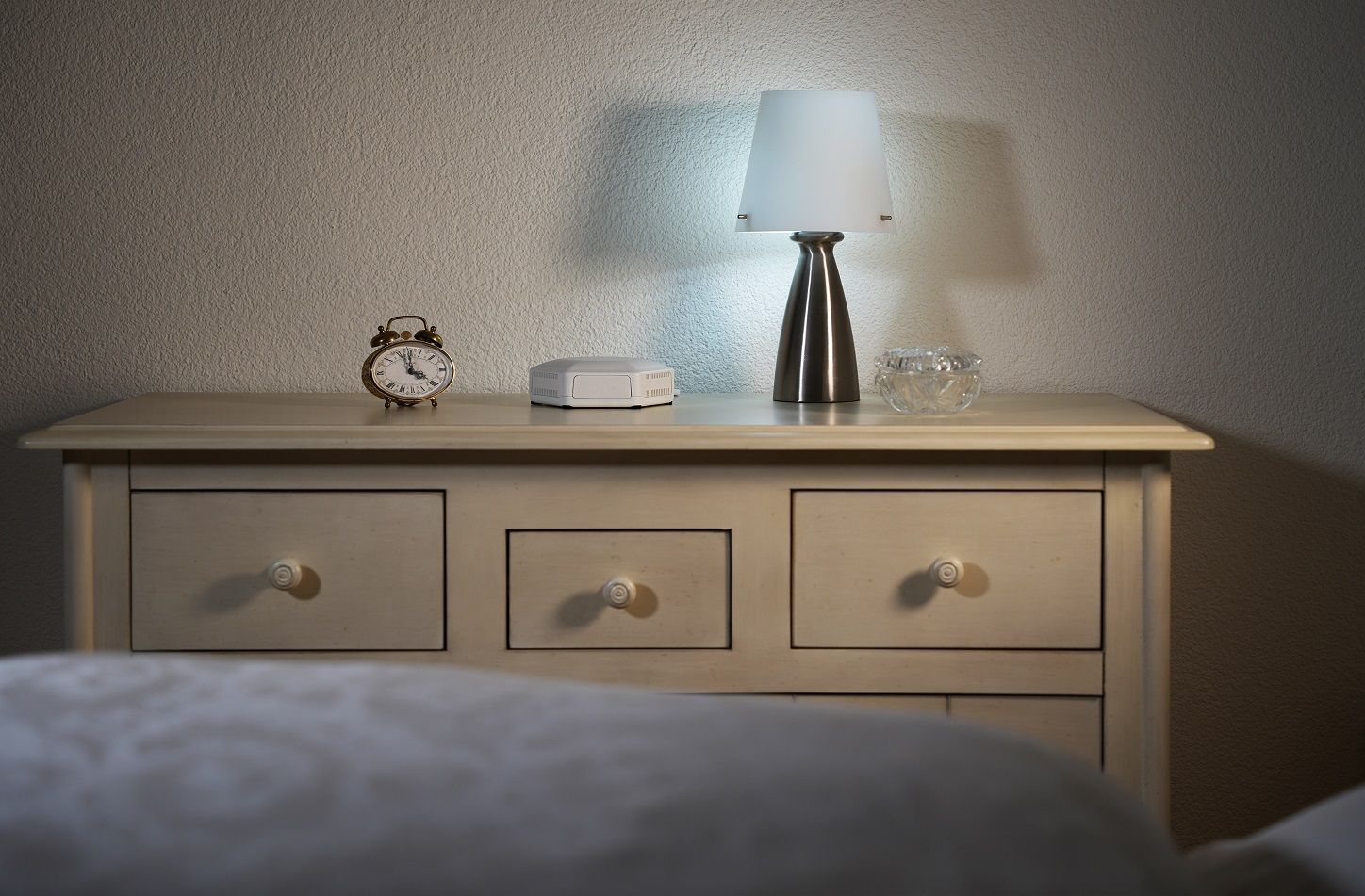 Wooden dresser with three drawers, an old-style brass alarm clock showing 12:57, a white electronic device, a silver lamp with white shade lit, and a glass container on top.