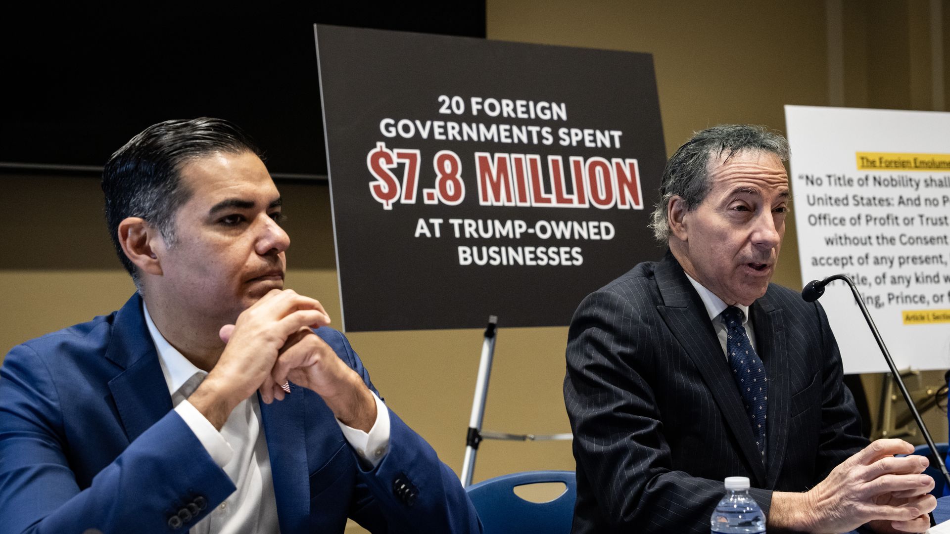 Two men in suits speak at a press event with a sign behind them reading "20 foreign governments spent $7.8 million at Trump-owned businesses." One man gestures while the other listens.