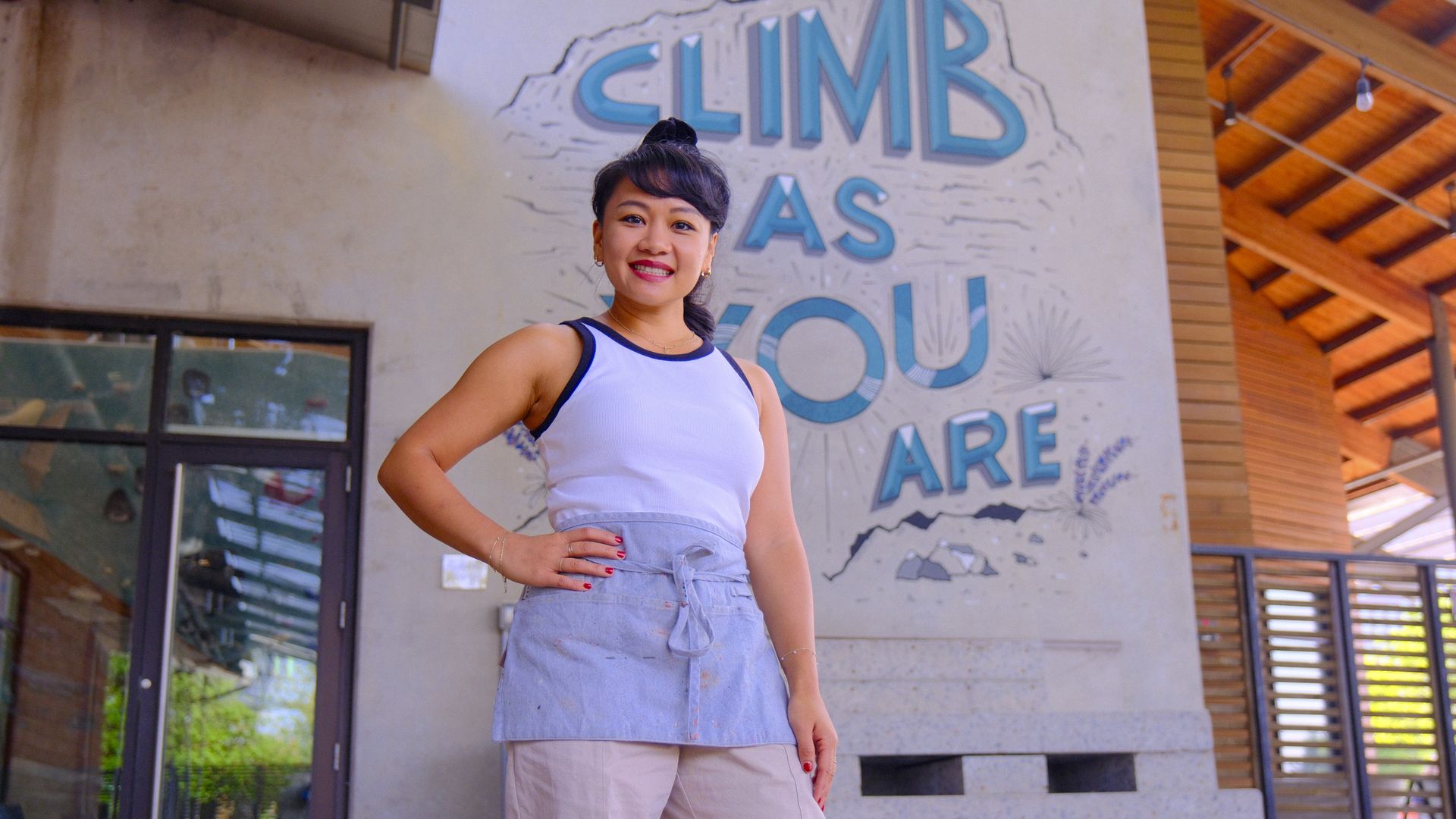 Dallas artist Jaclyn Le stands in front of a hand painted mural with the words climb as you are. 