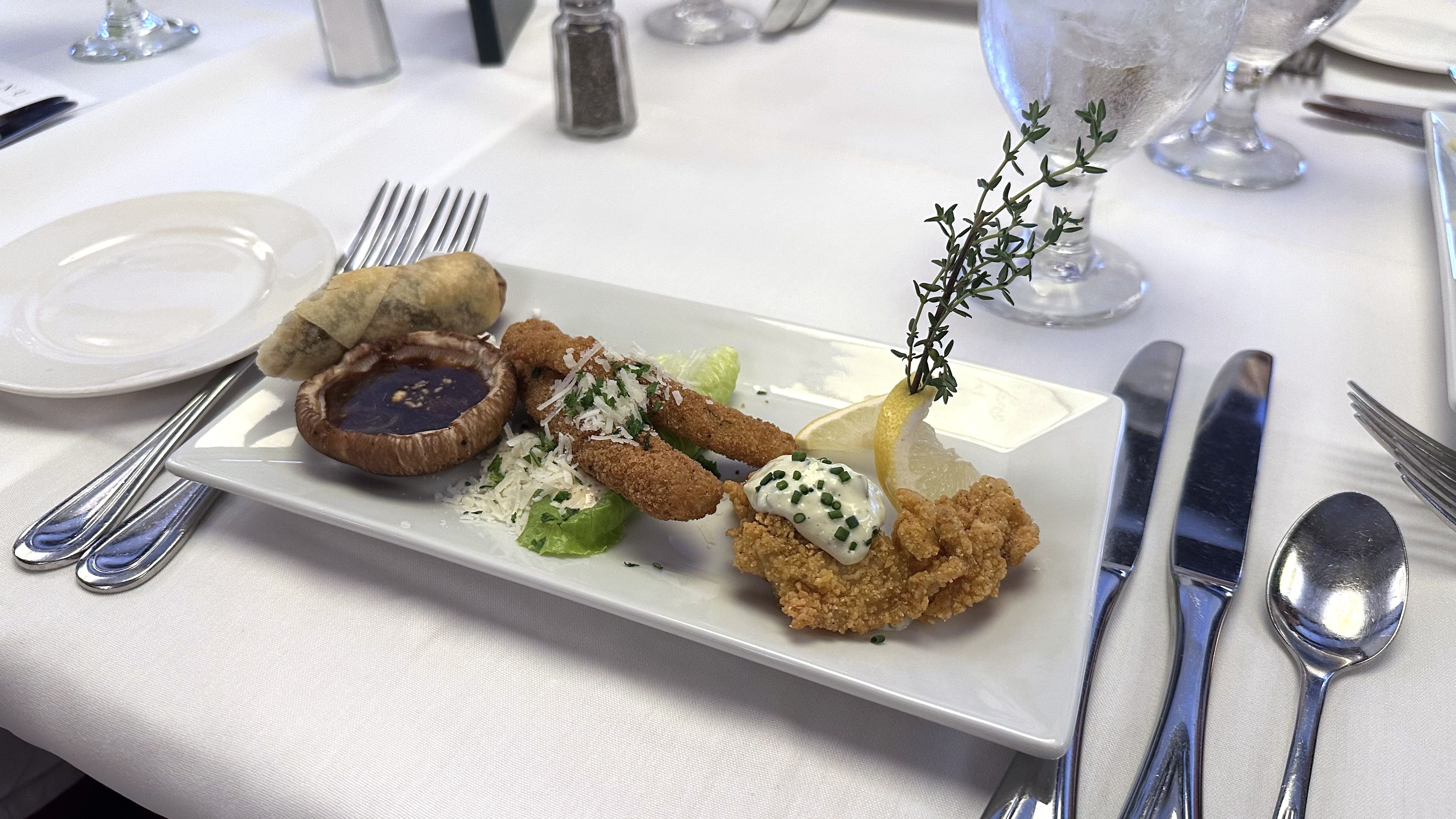 Rectangular white plate with assorted appetizers including a spring roll, sauce-filled mushroom, breaded sticks with grated cheese, and fried chicken with tartar sauce, garnished with lemon and herbs.