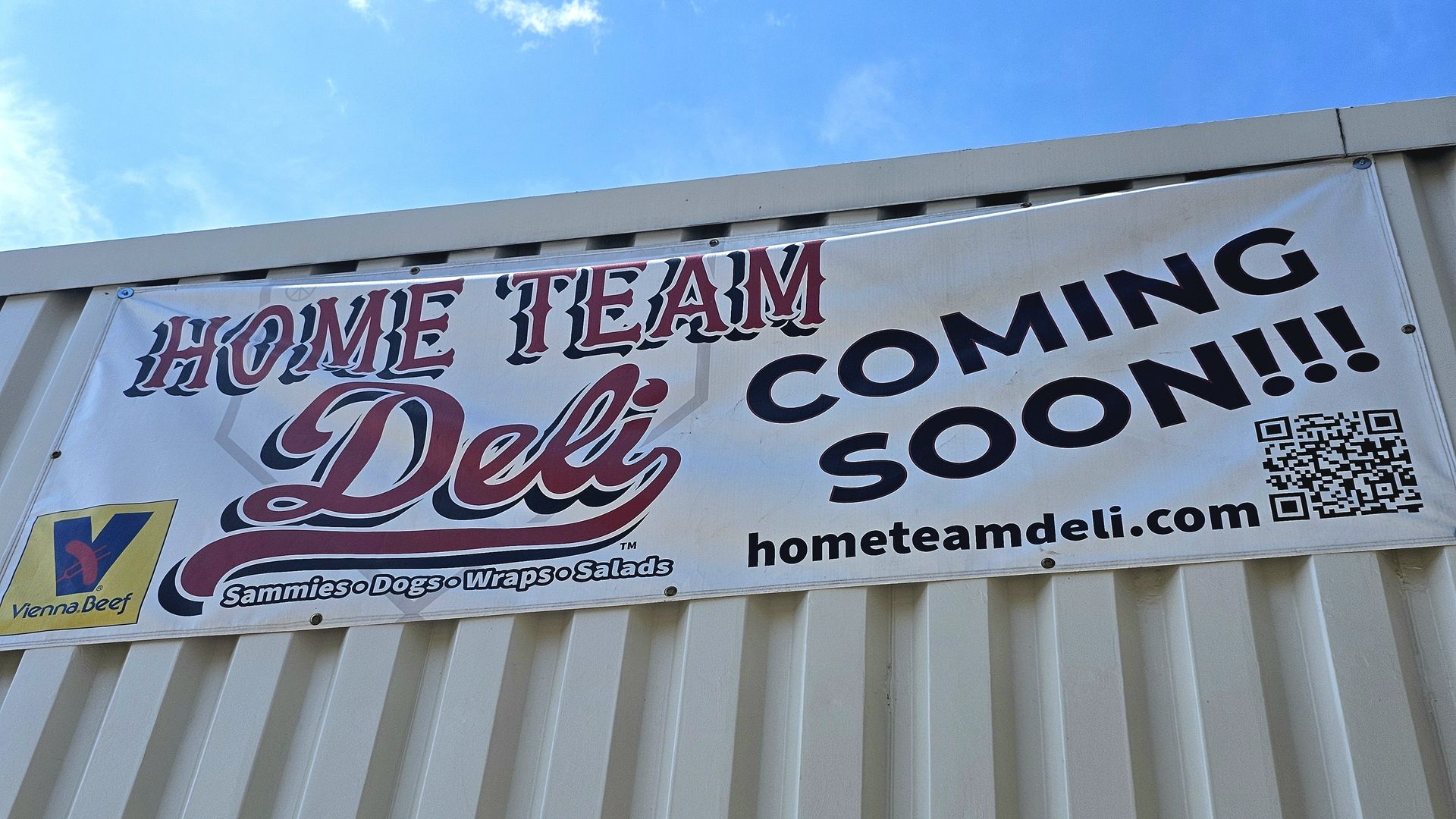 Banner on a light building displays 'HOME TEAM DELI' in red script and 'COMING SOON!!!' in bold black letters, with a QR code and 'hometeamdeli.com' under a blue sky.