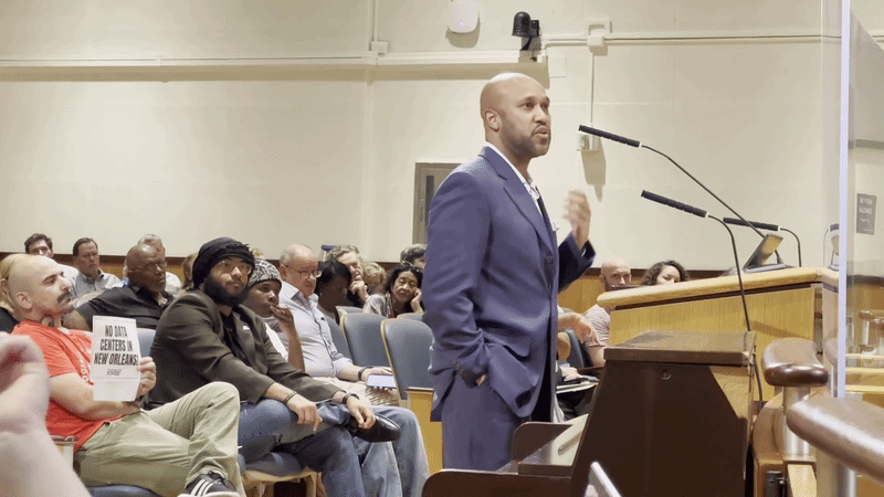 A man wearing a suit speaks at a podium in City Council chambers. Behind him in the crowd, a person holds a sign reading "No data centers in New Orleans."