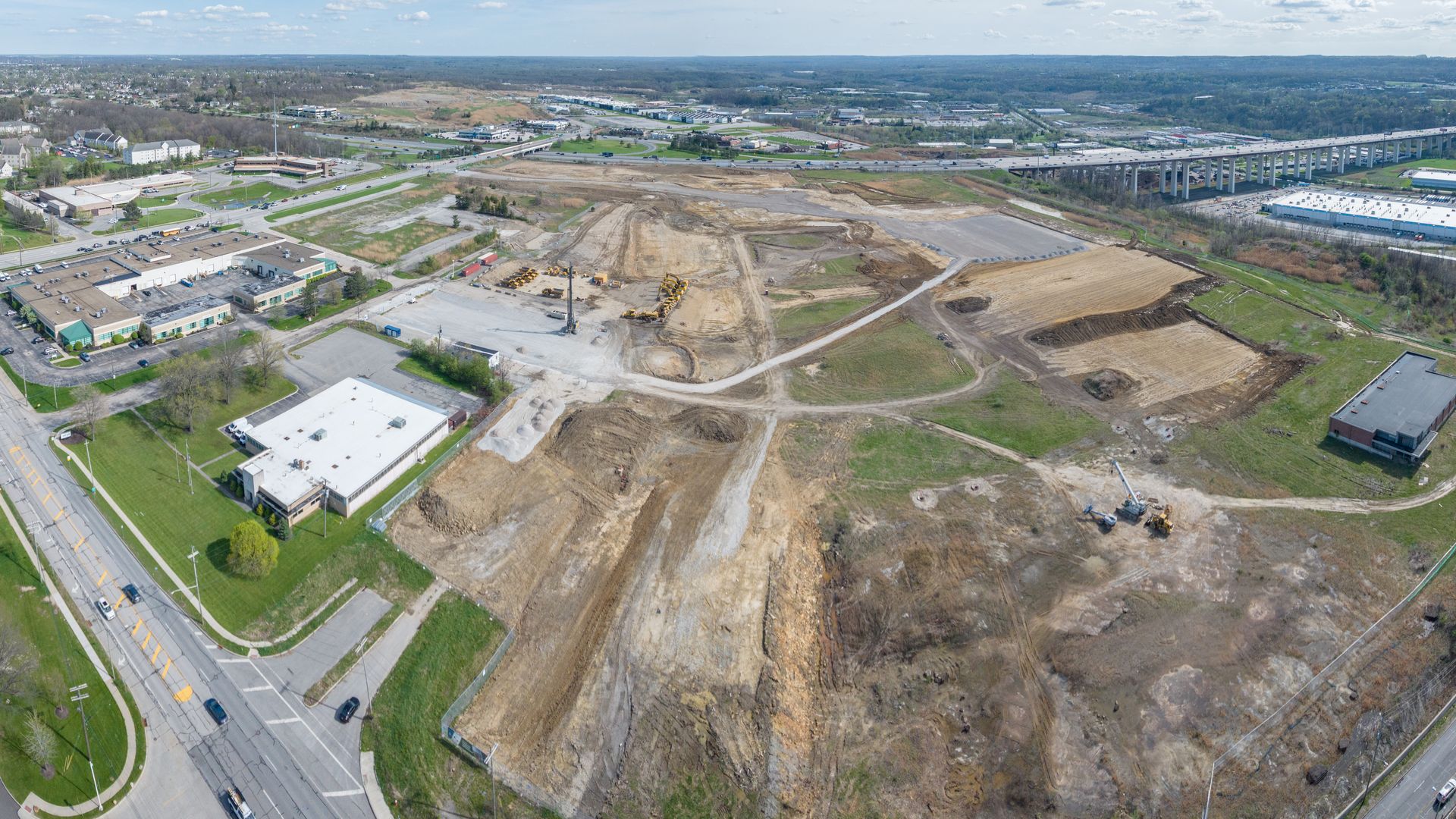 Aerial view of a town with a large construction site at center: dirt roads, earthworks, and excavators among soil mounds, surrounded by buildings, streets, and a long elevated highway on the right.