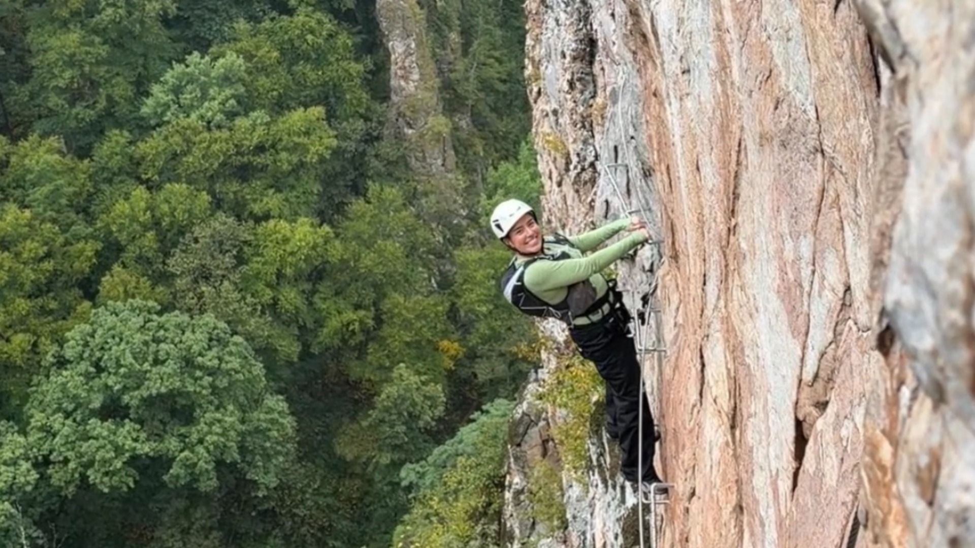 Climber in a white helmet, green shirt, and harness scales a rocky cliff, secured by rope, with dense green forest in the background.