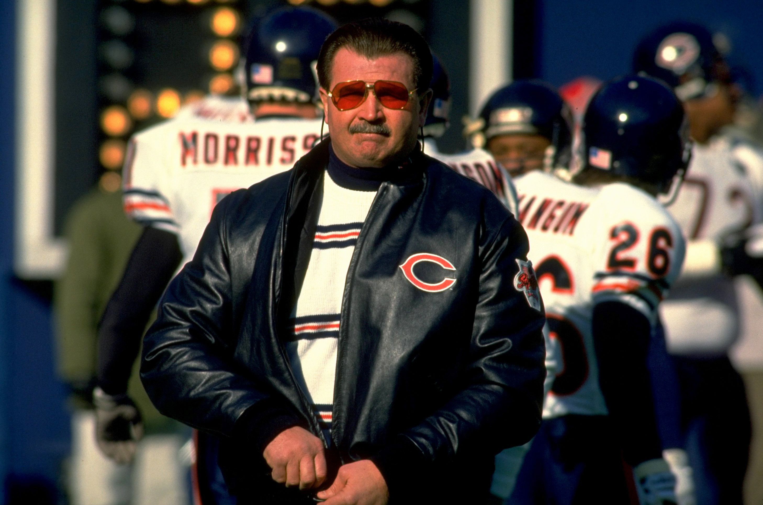 Photo of a man dressed in a leather jacket with sunglasses and a mustache walking on a football field
