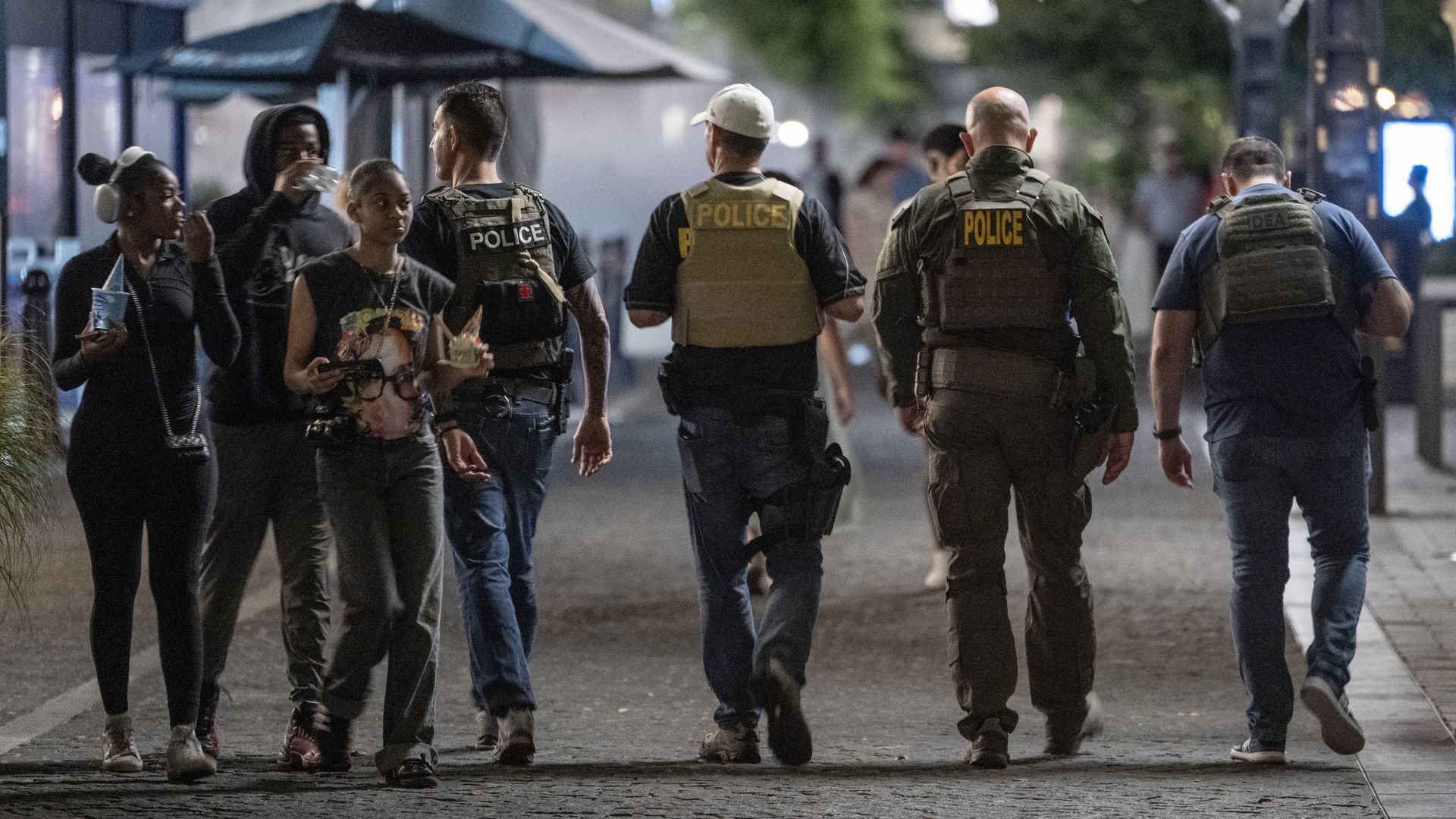 People walking on a city street at night, including four police officers wearing tactical vests with "POLICE" labels and three casually dressed individuals, some holding food and drinks.