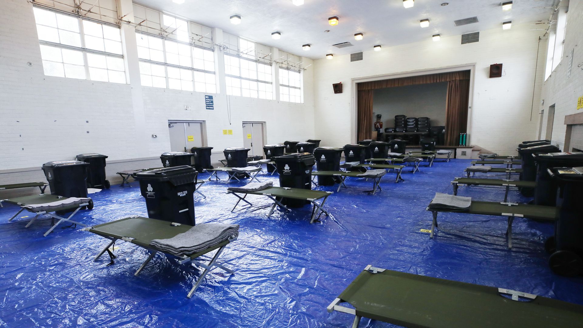 In this image, cots are lined up next to trash cans in a room lined with tarps.