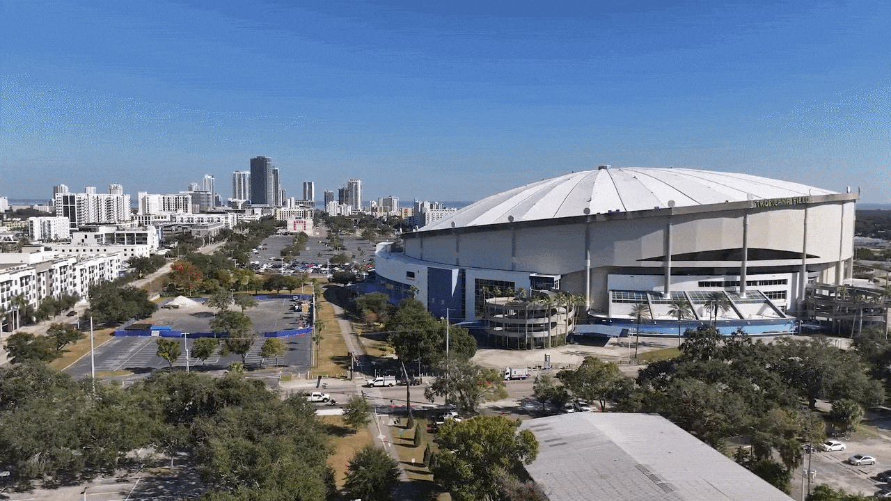 Aerial view of a large white-domed stadium with blue accents named Tropicana Field on the right, surrounded by parking lots, trees, and a distant city skyline under a clear blue sky.