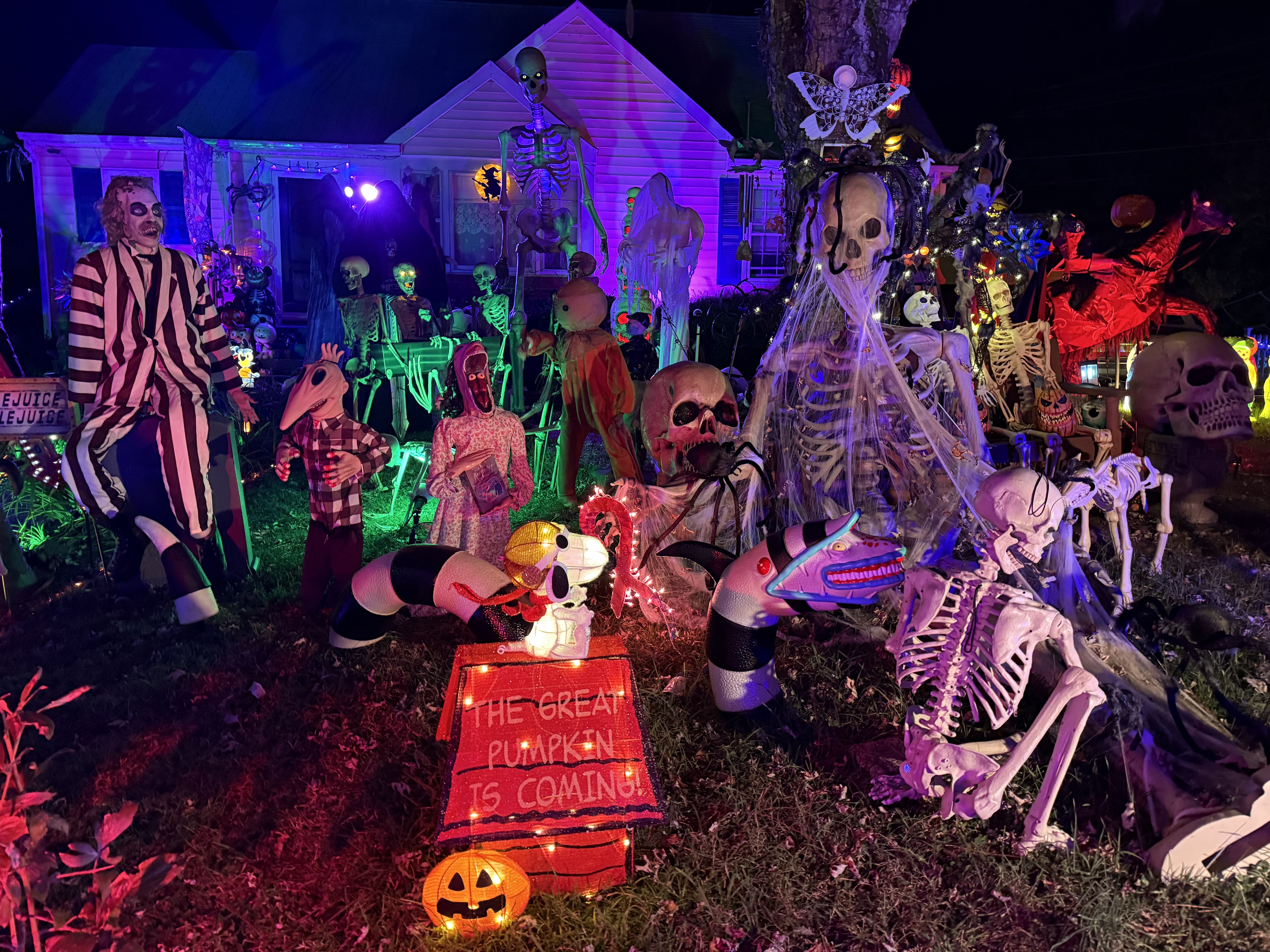 Nighttime Halloween yard display with colorful lights, dozens of skeletons, ghosts, pumpkins, and eerie figures in a front yard; a striped person stands left, and a sign reads "THE GREAT PUMPKIN IS COMING" near the ground.