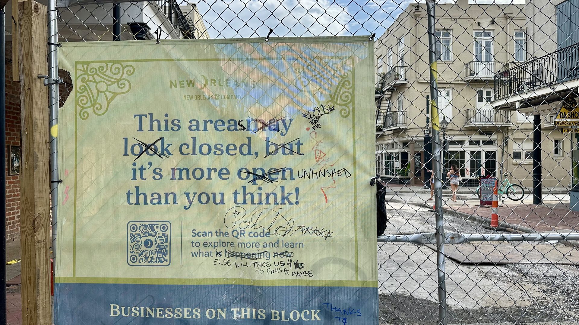 Construction banner behind a chain-link fence. It reads, 'This area may look closed, but it's open' with a green decorative border. In the background are beige buildings, a person, and a bicycle.